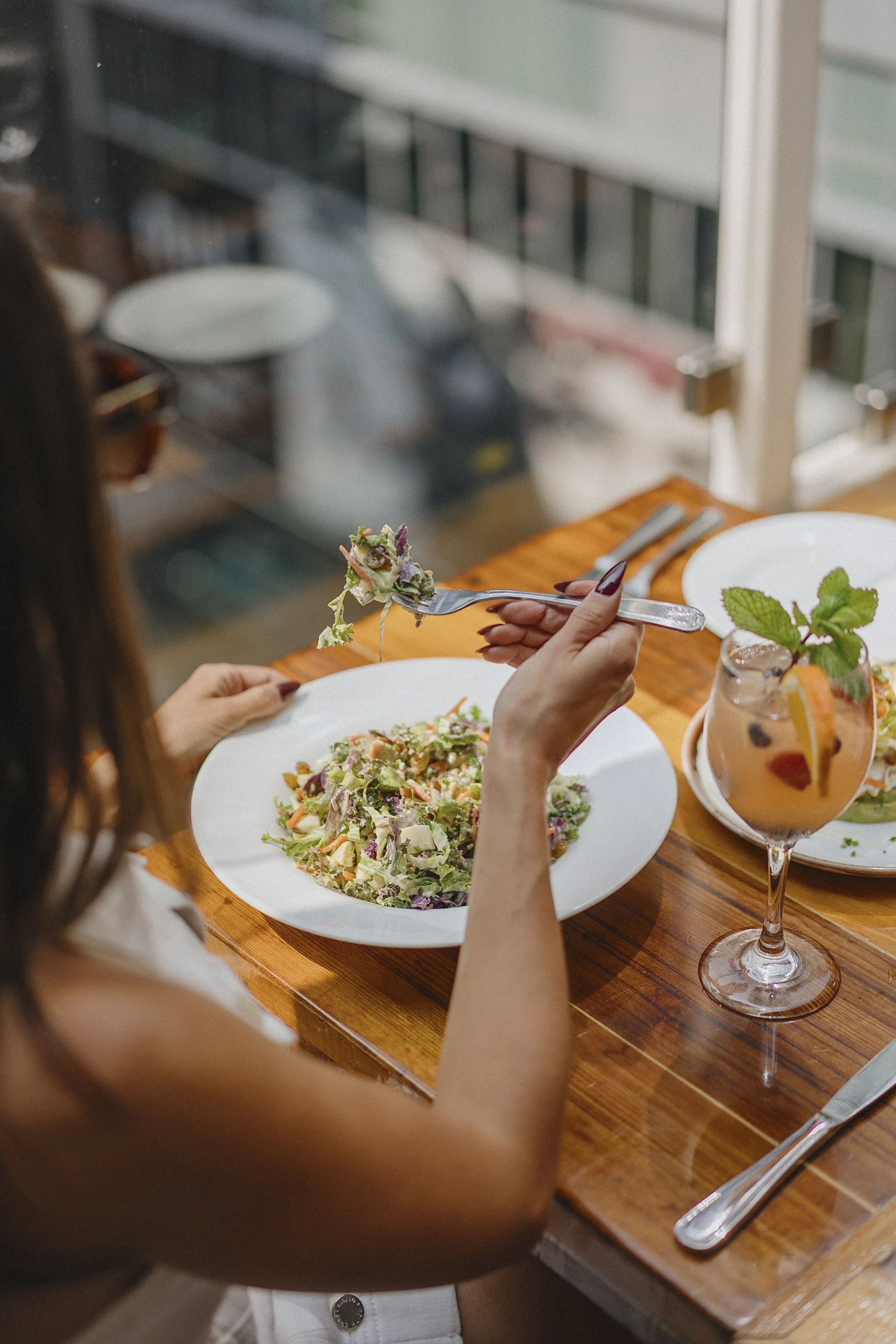 A woman holding a fork with a bite of salad, sitting at a wooden table with a large salad plate and a cocktail garnished with mint and a slice of orange.