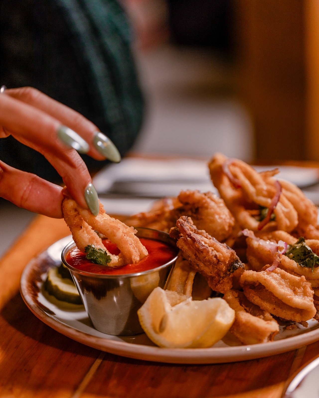 Person dipping fried onion rings into a small bowl of ketchup at a restaurant. The plate includes more fried onion rings, lemon wedges, and dipping sauces.