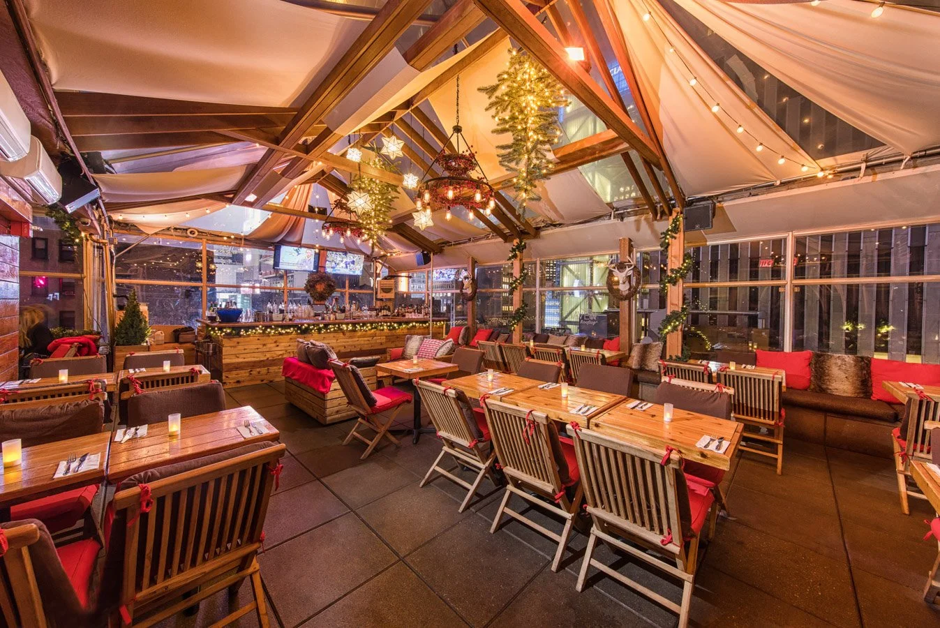 Indoor restaurant decorated for Christmas with wooden tables, chairs with red cushions, garlands, Christmas lights, and a bar area in the background.