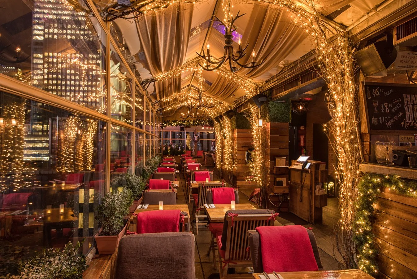 A cozy restaurant or bar interior decorated for the holidays with string lights, fairy lights on wooden beams, and red and gray chairs around wooden tables set with candles.