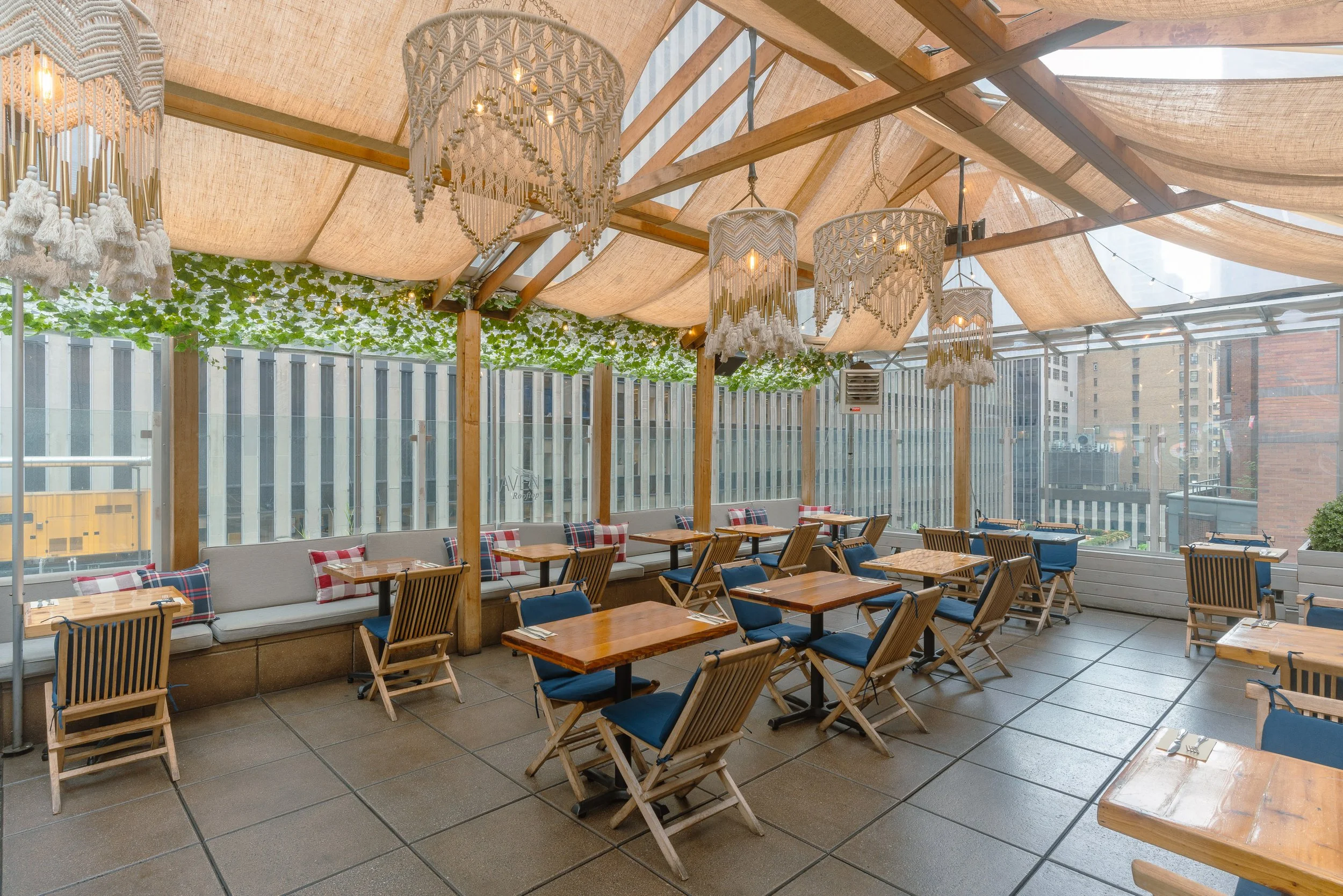 Interior of a restaurant or cafe with wooden tables and chairs, decorated with plaid pillows, large macramé chandeliers hanging from a wooden ceiling, and a wall covered with greenery and string lights.