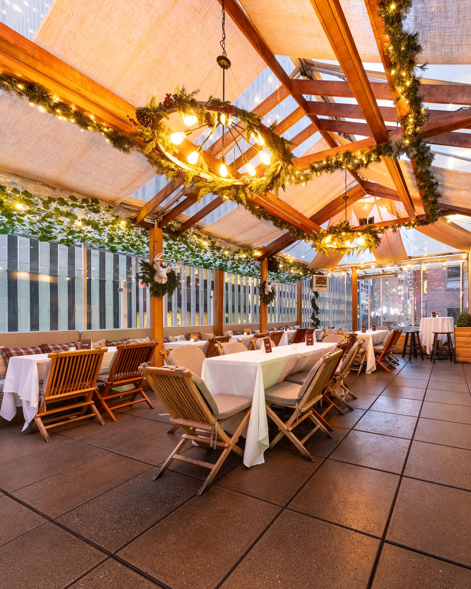A decorated restaurant seating area with white tablecloths, wooden chairs, and festive greenery with string lights and garlands hanging from the ceiling beams.