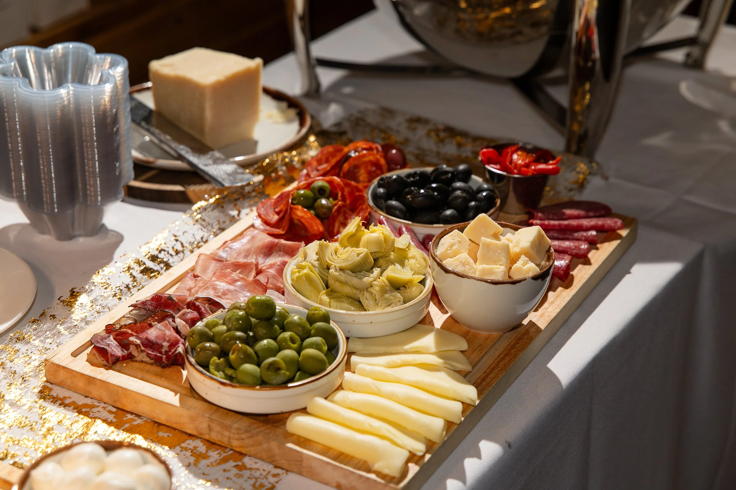 A charcuterie board with various cheeses, meats, olives, artichoke hearts, and peppers on a wooden cutting board, decorated with gold confetti.