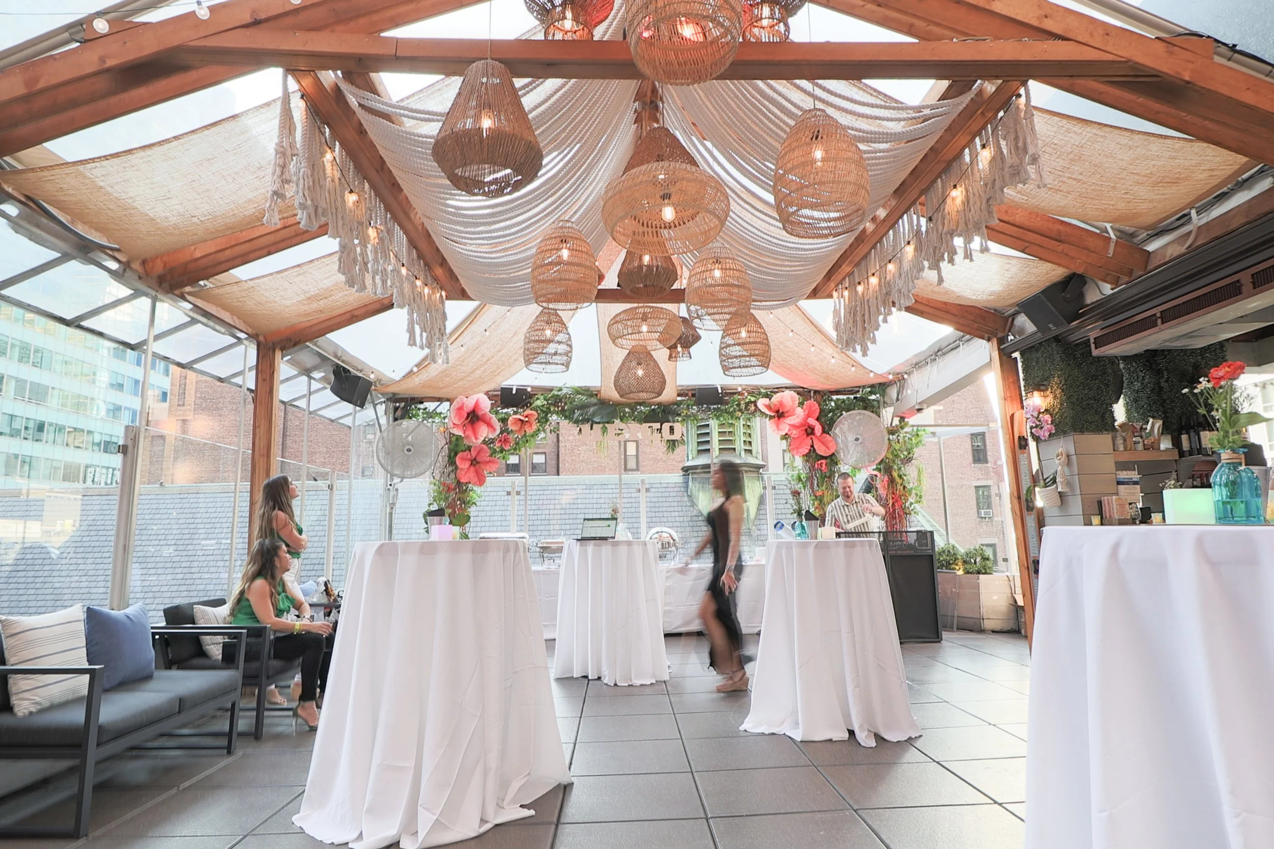 Indoor event space decorated with hanging wicker and fabric lamps, string lights, pink flowers, and white drapes. There are tables covered with white cloths and a few people seated and standing.