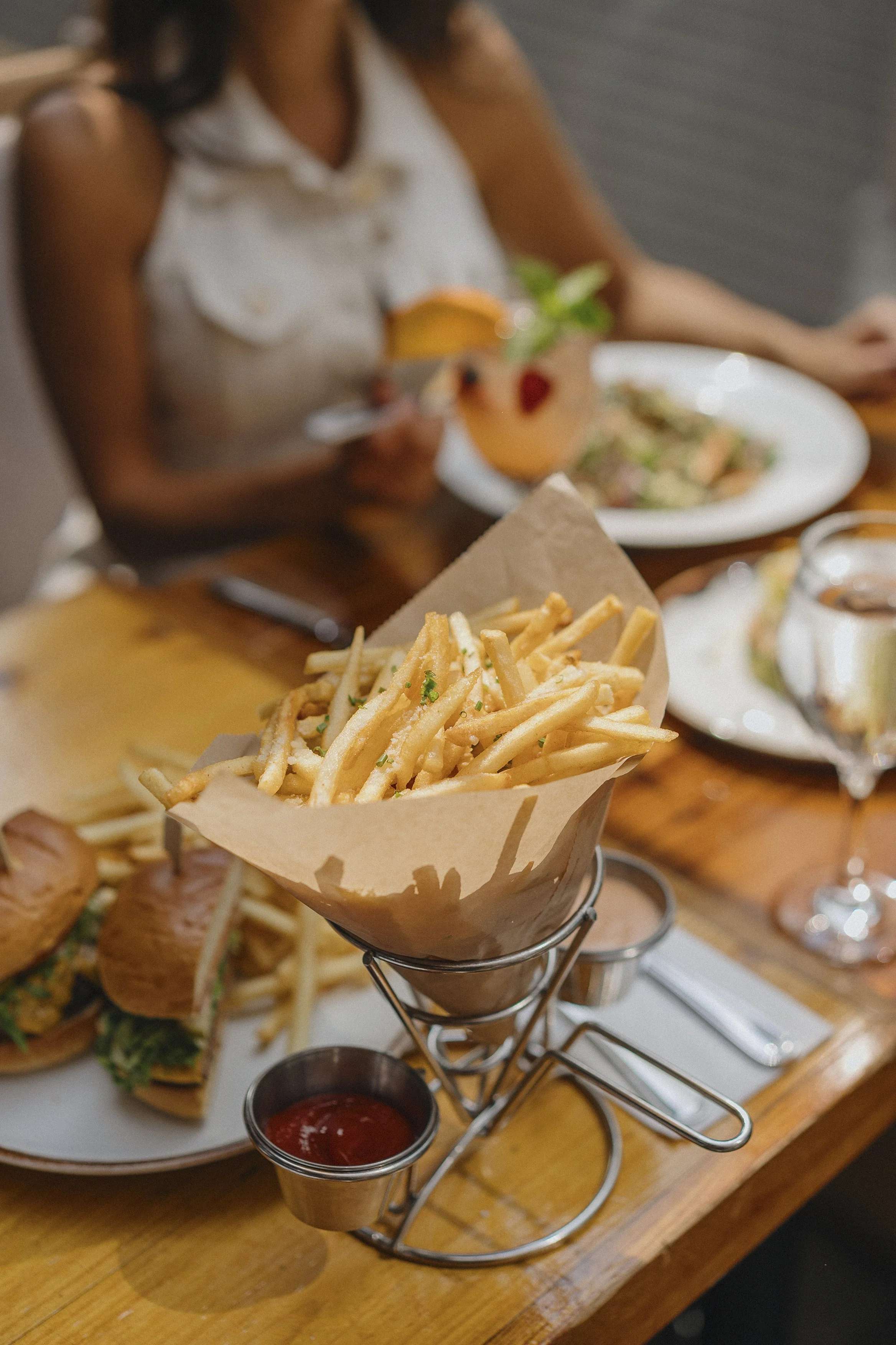 Fried French fries served in a paper cone on a metal basket stand, with a small cup of ketchup on a wooden table. In the background, there are plates of food and a woman holding a cocktail drink garnished with citrus and mint, at a restaurant or cafe