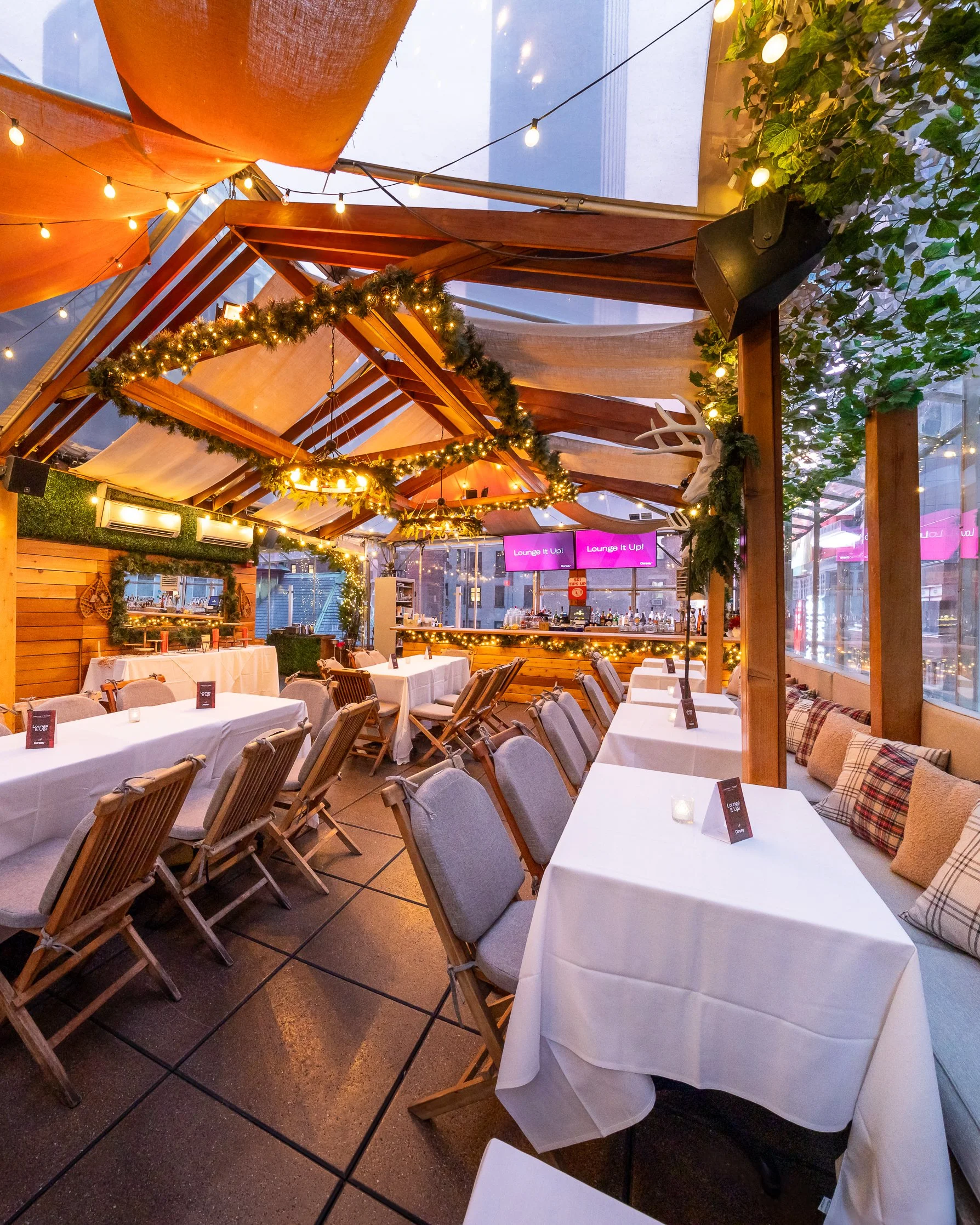 Indoor restaurant or event space with tables covered in white tablecloths, chairs, and decorative greenery. String lights hang from the ceiling, and a bar area is visible in the background. A chandelier with greenery surrounds it, and a mounted white