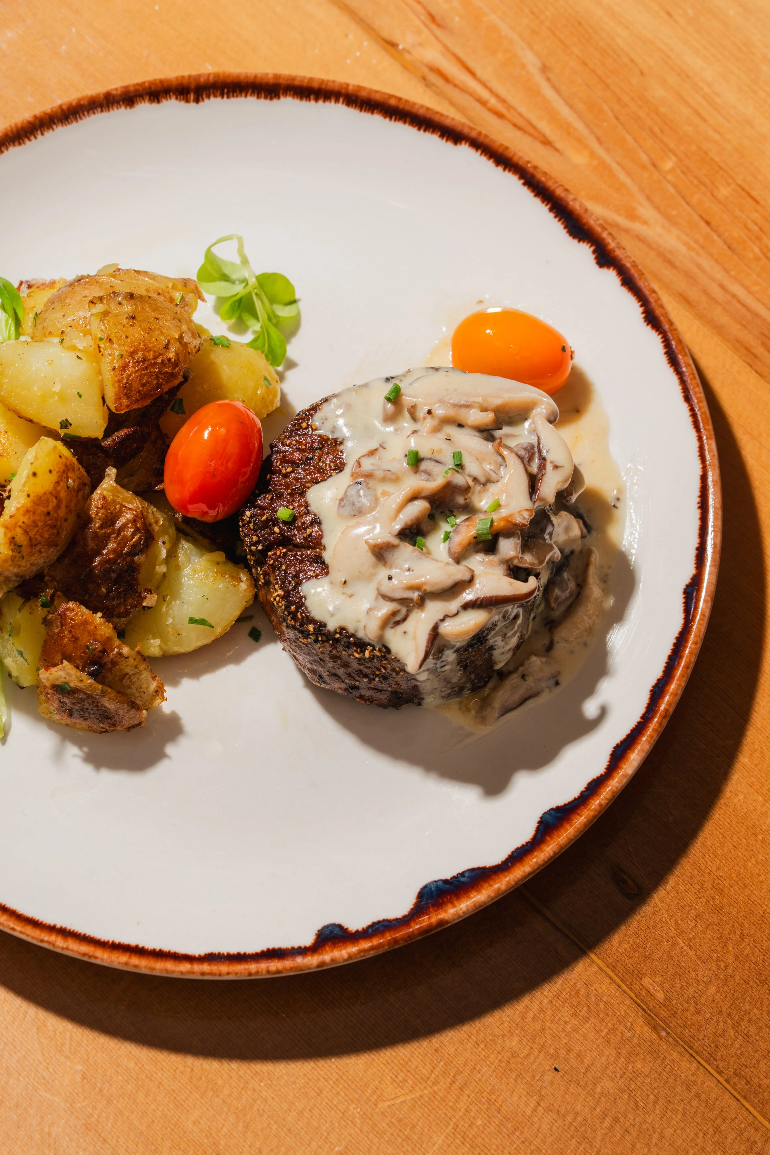 Plate with roasted potatoes, cherry tomatoes, and a beef steak topped with mushroom sauce, garnished with chives, on a wooden table.