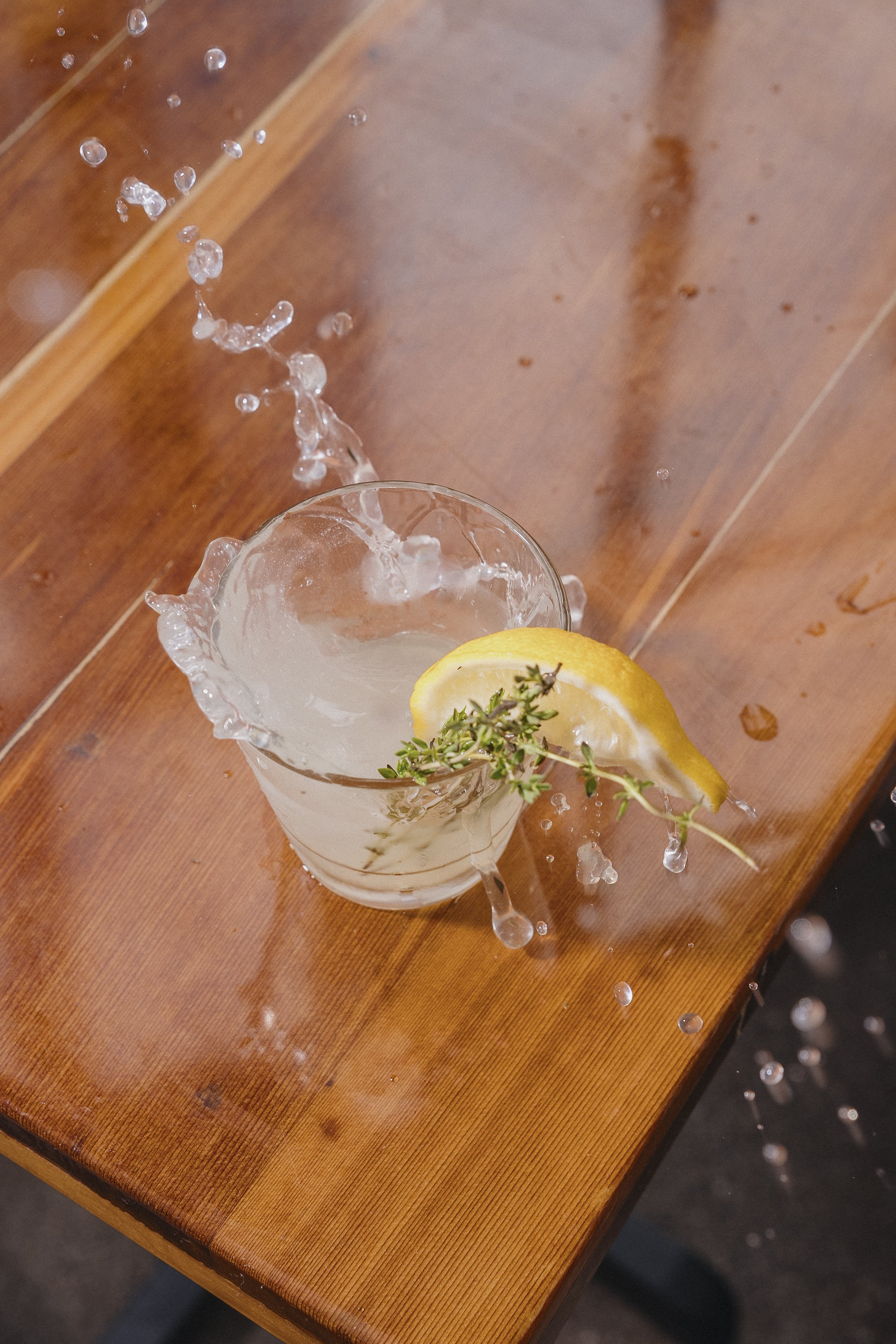 A wooden table with a glass containing a lemon slice, sprigs of herbs, and ice, with water splashing out of the glass.