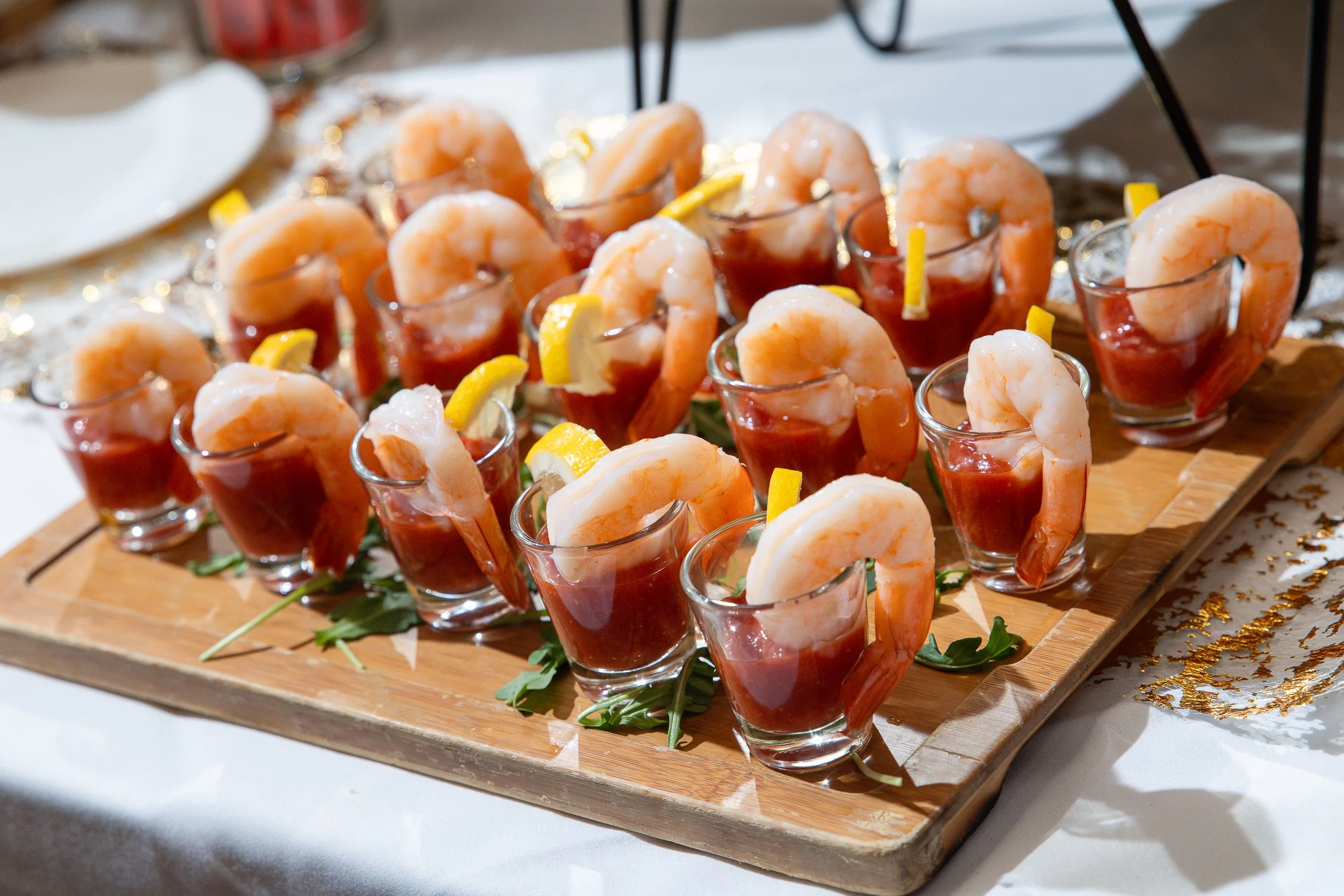 A display of shrimp cocktails served in small glasses with lemon wedges, arranged on a wooden serving board at a party or event.