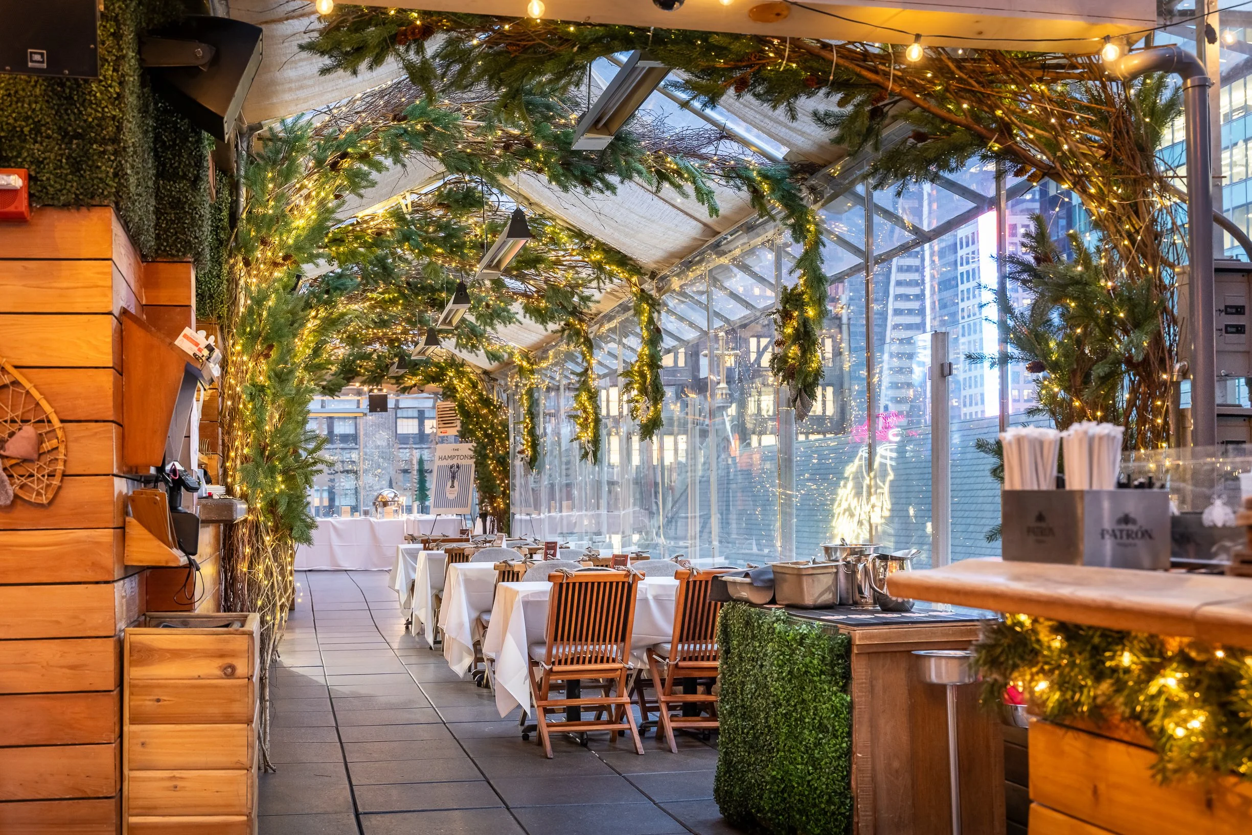 Indoor restaurant decorated with Christmas greenery and string lights, set up with tables and chairs, with city skyscrapers visible outside.