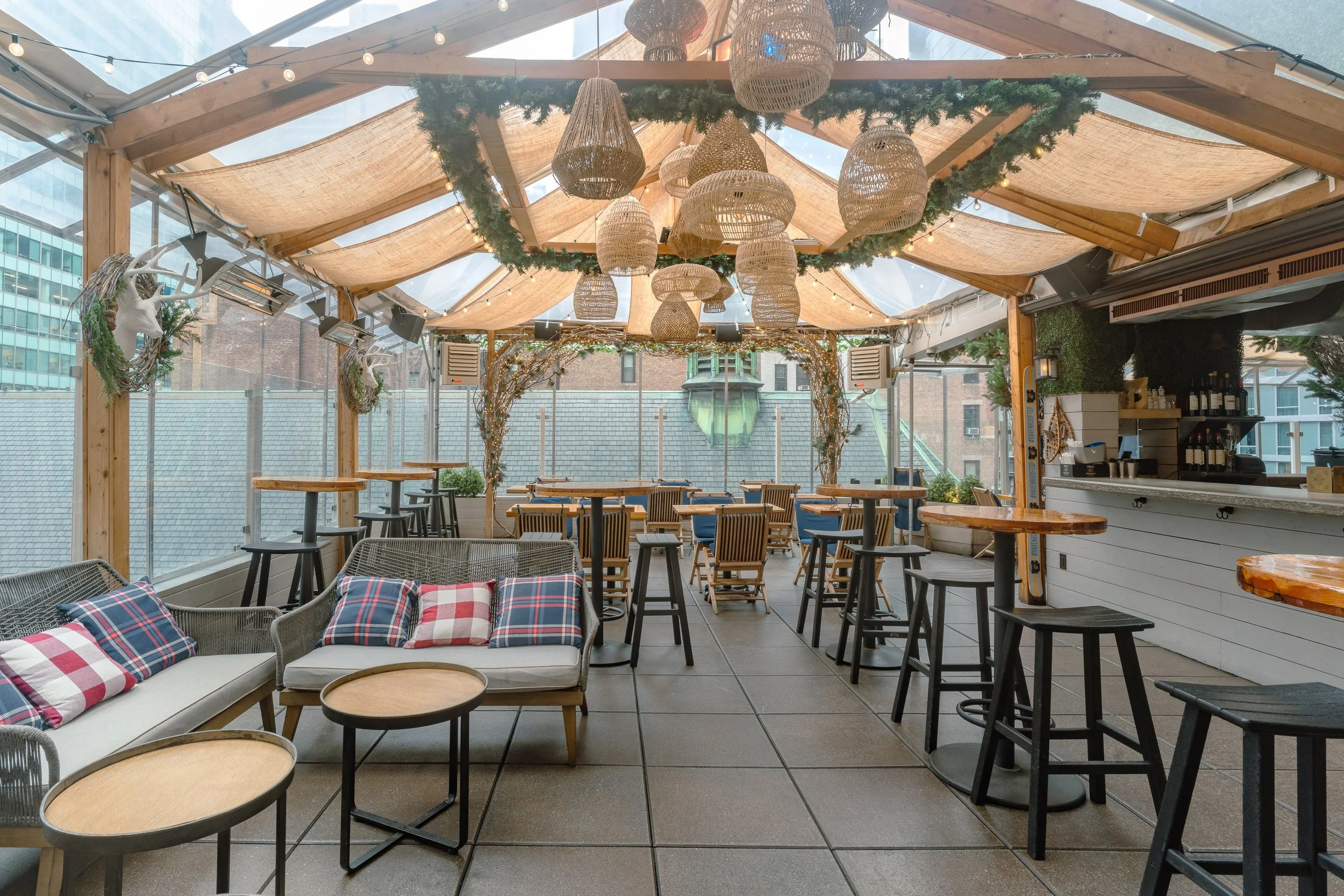 Decorative outdoor restaurant or café with wooden beams, wicker pendant lights, and holiday greenery. There are tables and chairs, including a white sofa with plaid pillows, and a bar area visible on the right. A glass roof allows natural light.