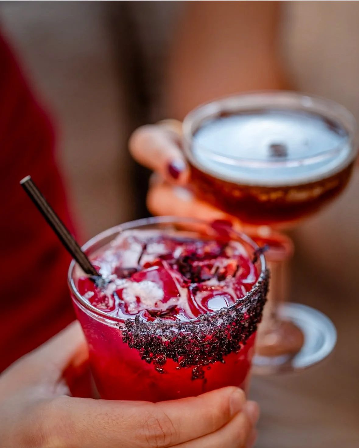 Person holding a glass with a red cocktail garnished with fruit, with a second person holding a glass of red wine in the background.