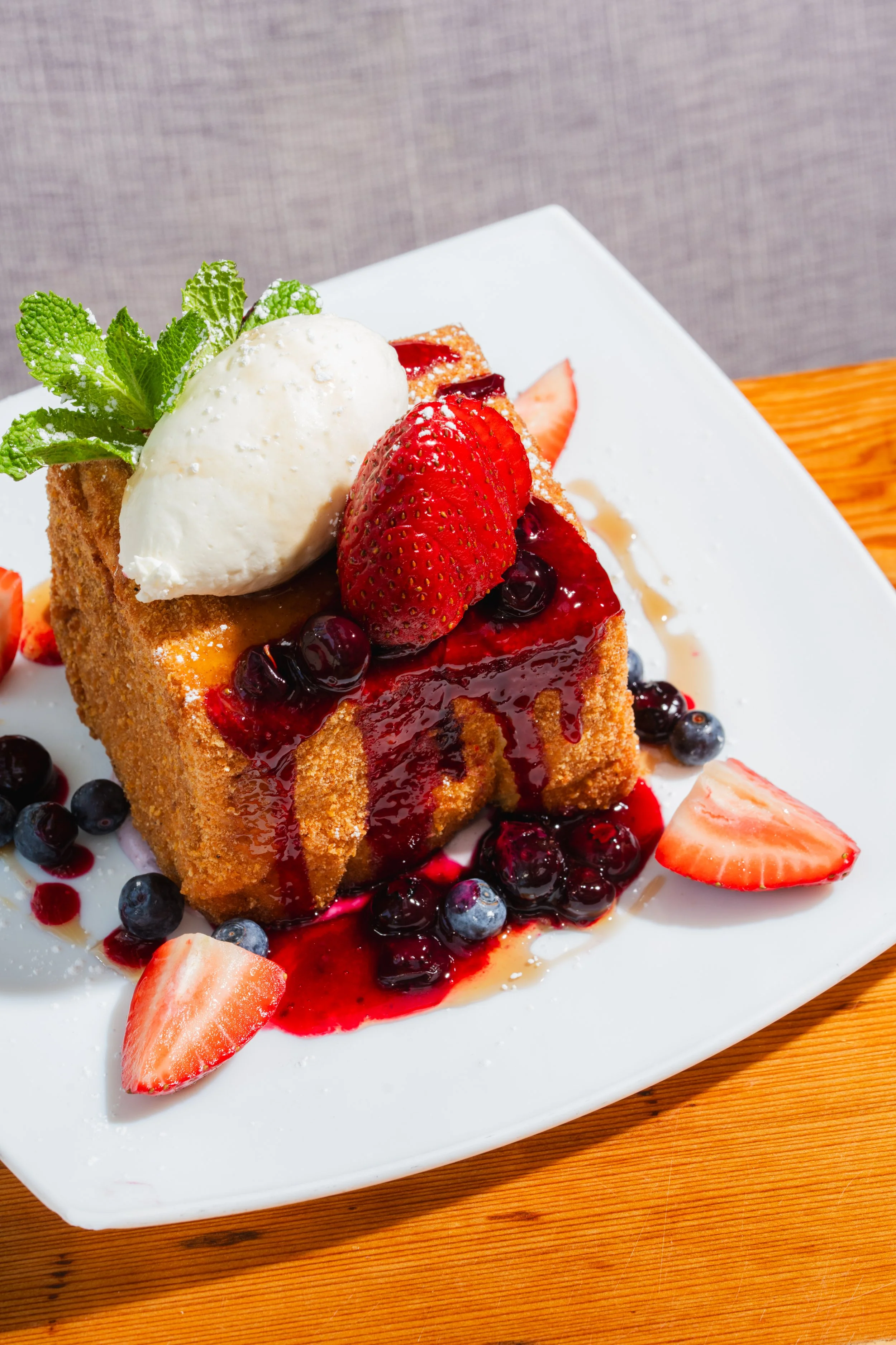 A dessert with a slice of cake topped with strawberries, blueberries, a scoop of ice cream, and mint leaves, served on a white plate.