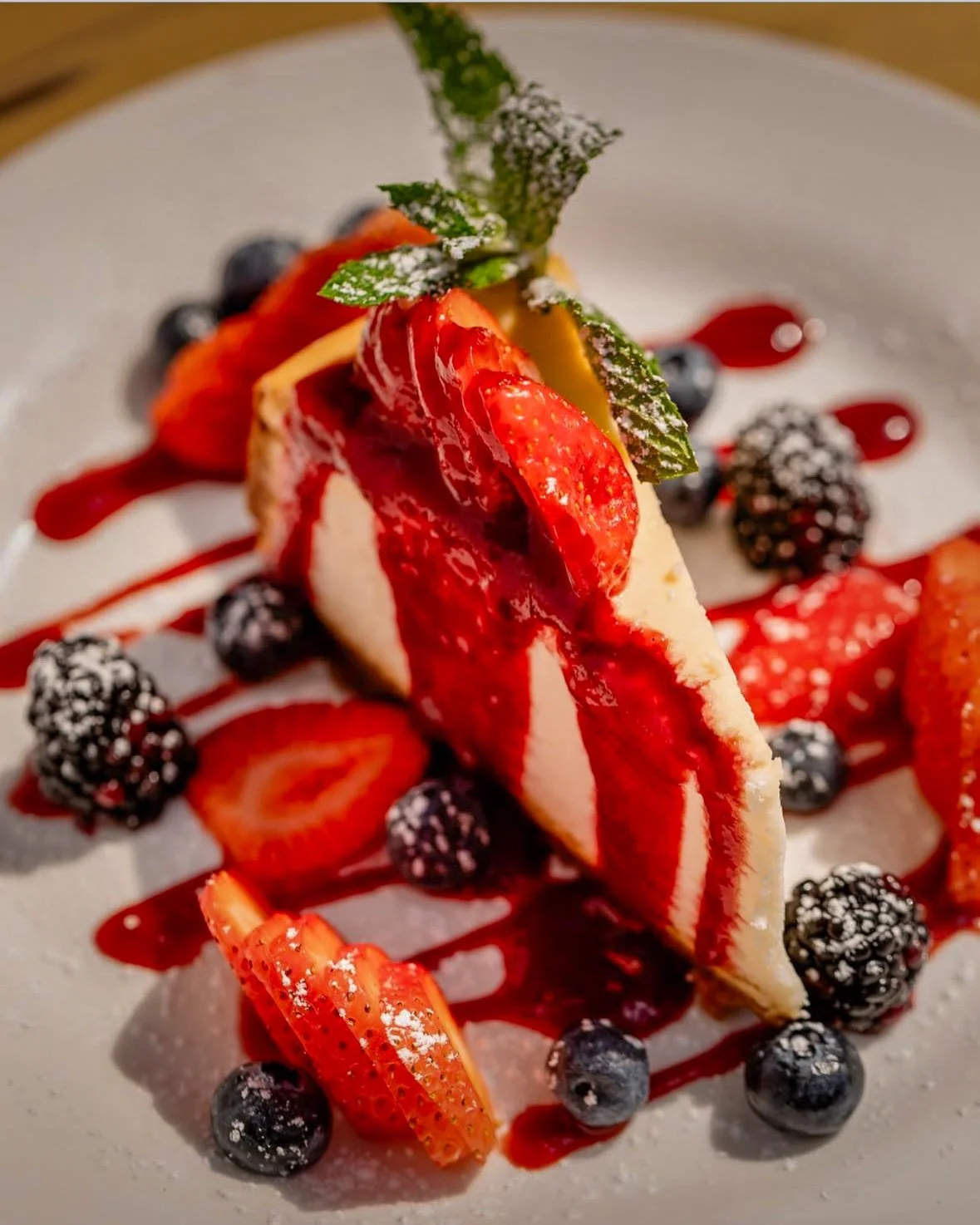 Slice of cheesecake topped with strawberries, blueberries, and mint leaves, drizzled with berry sauce, served with additional berries and sliced strawberries on the side.
