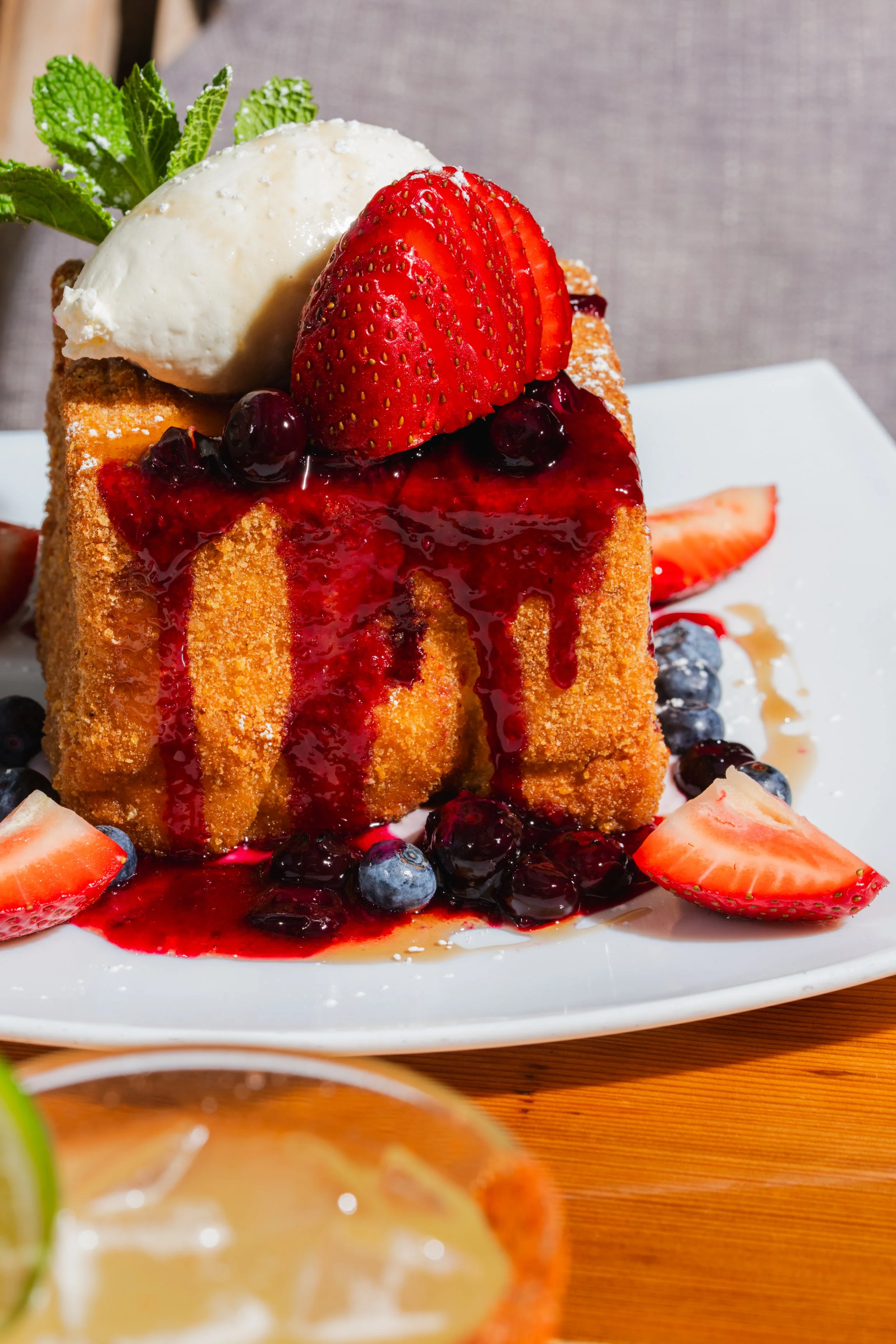 Close-up of a dessert on a white plate, topped with whipped cream, sliced strawberries, and blueberries, with berry sauce drizzled over it.