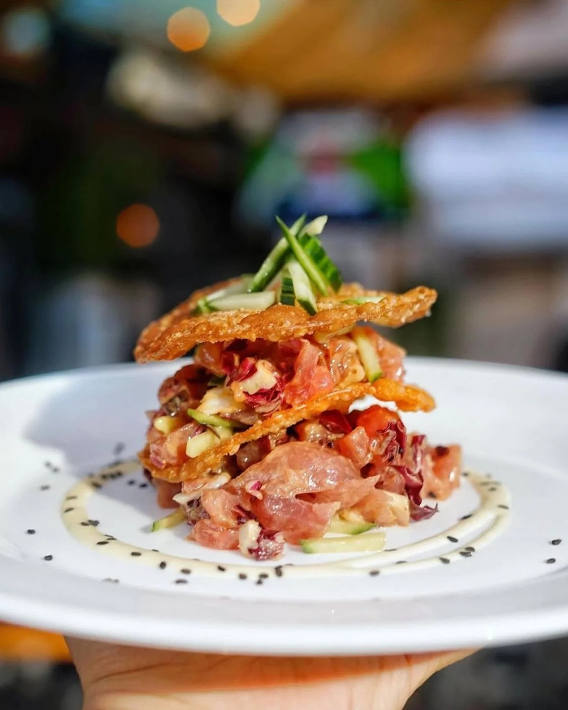 Close-up of a gourmet appetizer with layers of chopped vegetables and meat, topped with crispy chips and green garnishes on a white plate decorated with black sesame seeds.