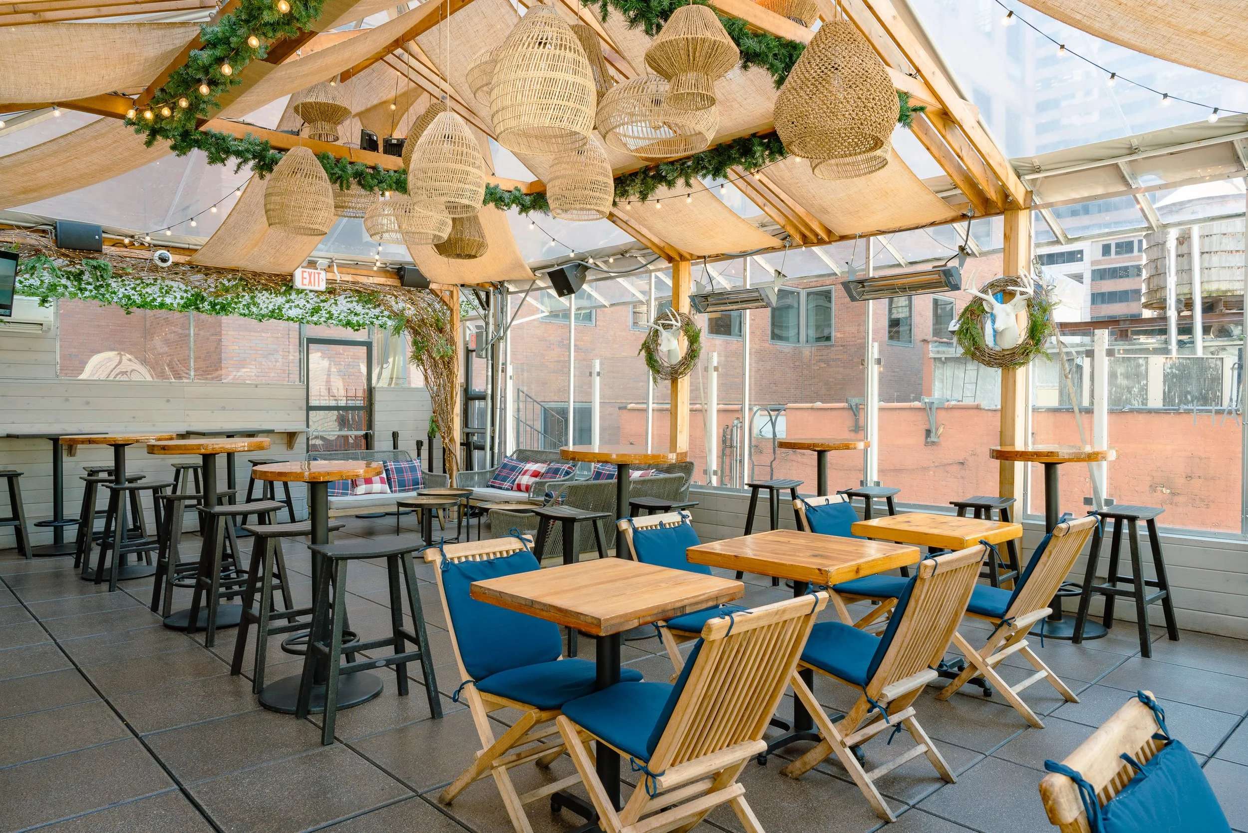 Indoor restaurant or café with wooden tables and blue cushioned chairs. Decor includes woven pendant lights, string lights, and green garlands with pine wreaths and reindeer heads on the walls. Large windows reveal an urban cityscape with brick and g
