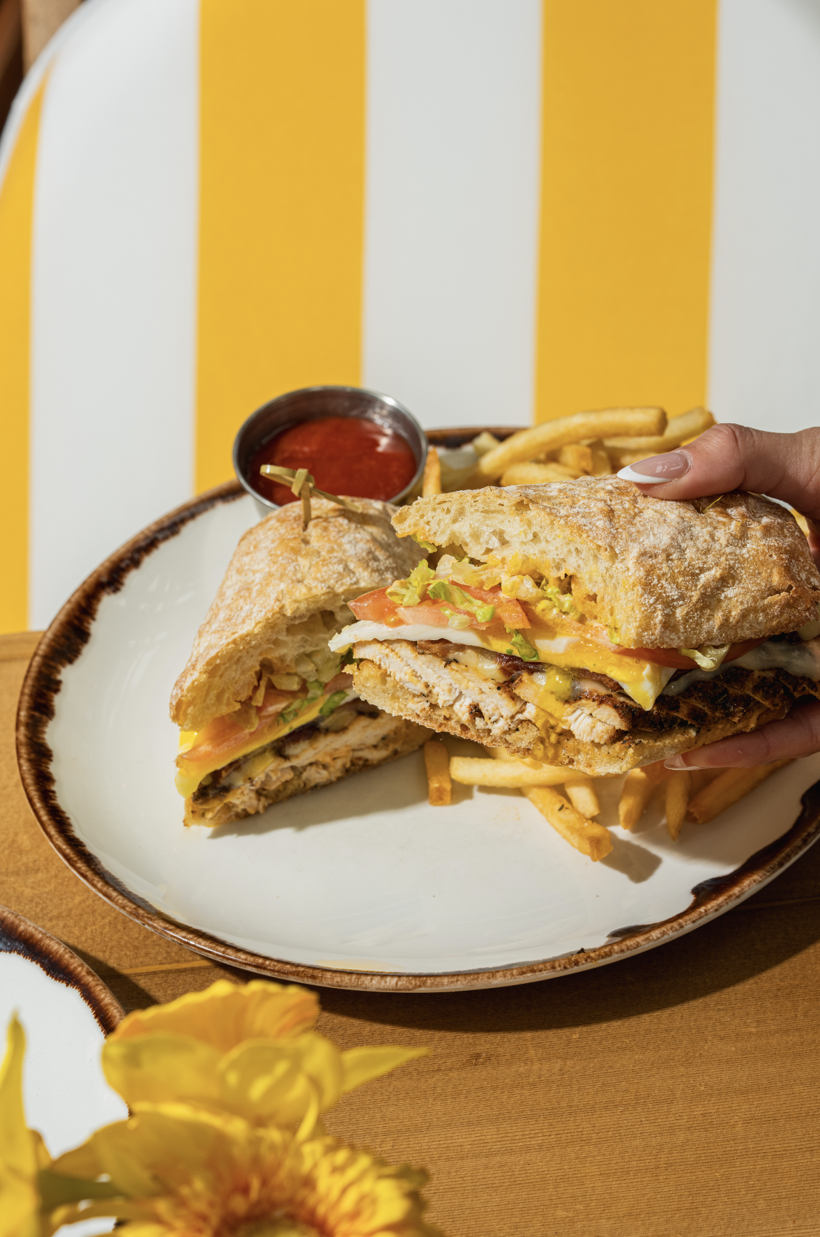 A handheld sandwich on a white and brown plate, with grilled chicken, lettuce, tomato, cheese, and mayonnaise, cut in half. There are French fries and ketchup in a small cup on the plate. Yellow flowers in the foreground.