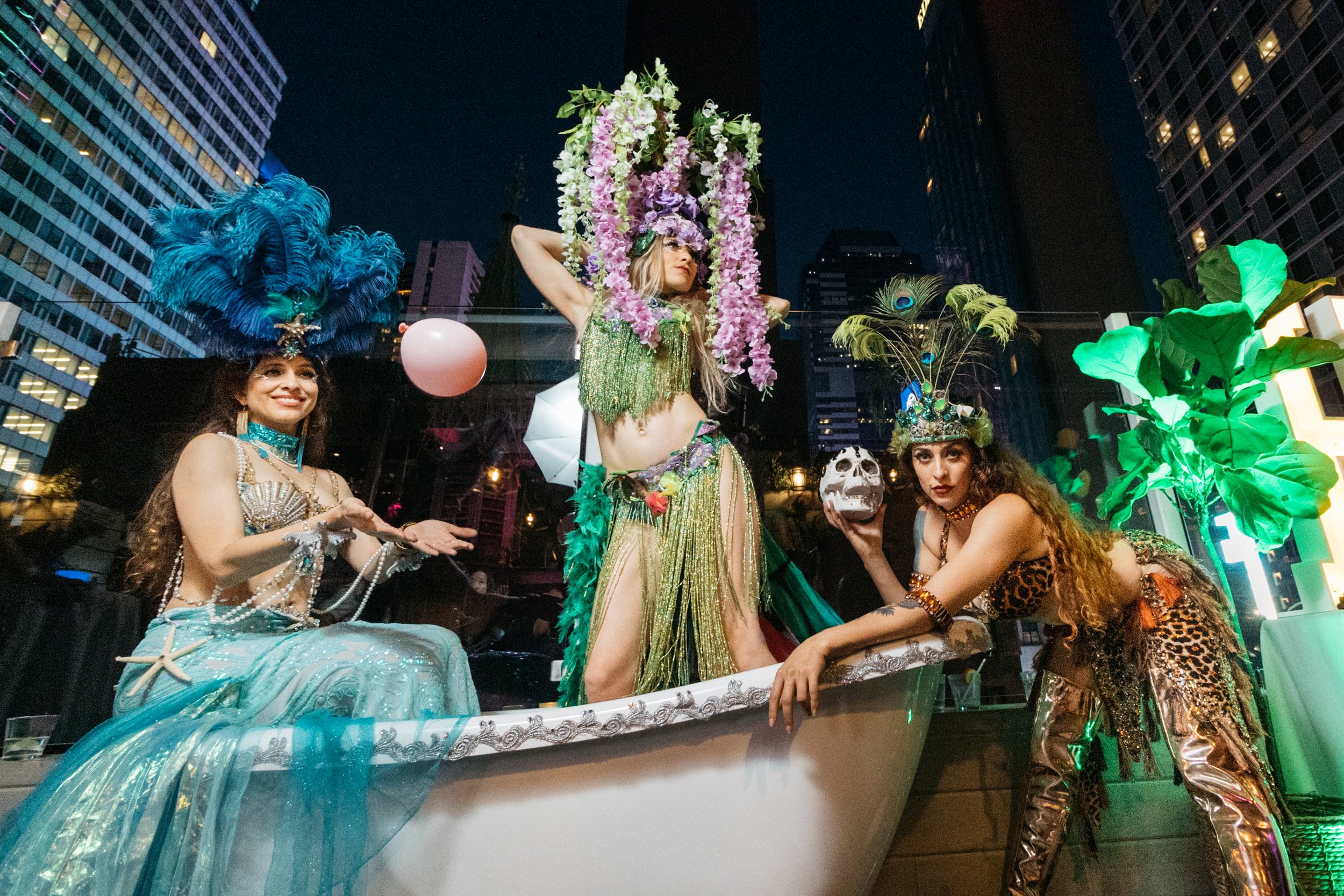 Group of women in elaborate costumes with tropical and ocean themes, on a parade float at night in an urban setting.