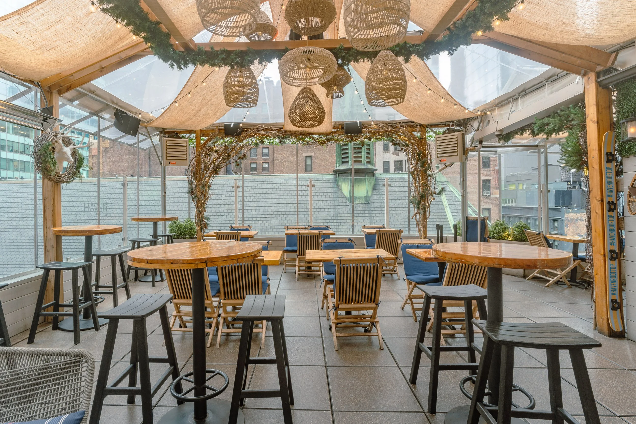 Indoor rooftop restaurant or bar with wooden tables and chairs, decorated with hanging wicker lamps, string lights, and greenery.