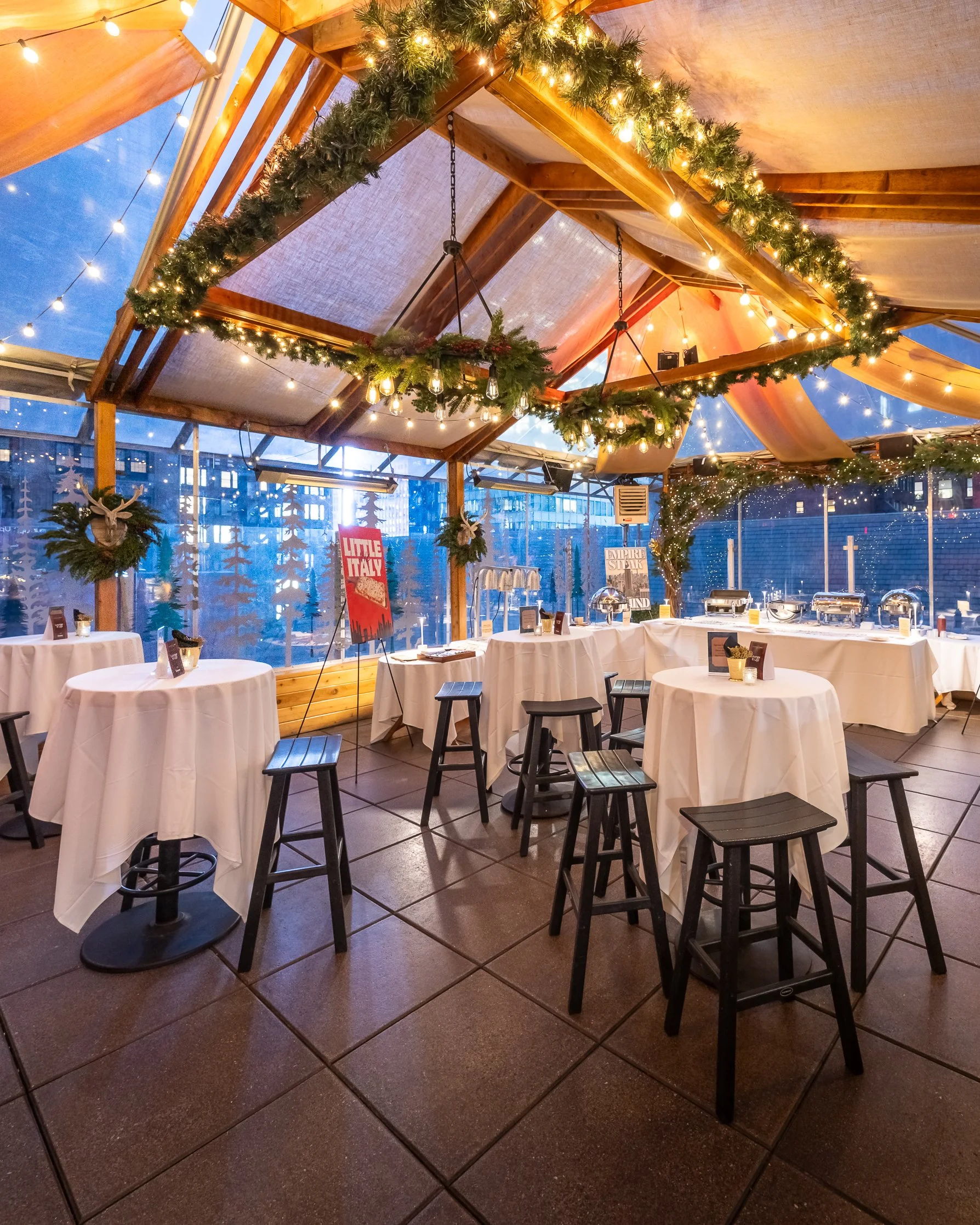 Indoor restaurant or banquet facility decorated for winter or Christmas with string lights and greenery, round tables with white tablecloths and black stools, wooden beams and large windows showing a cityscape.