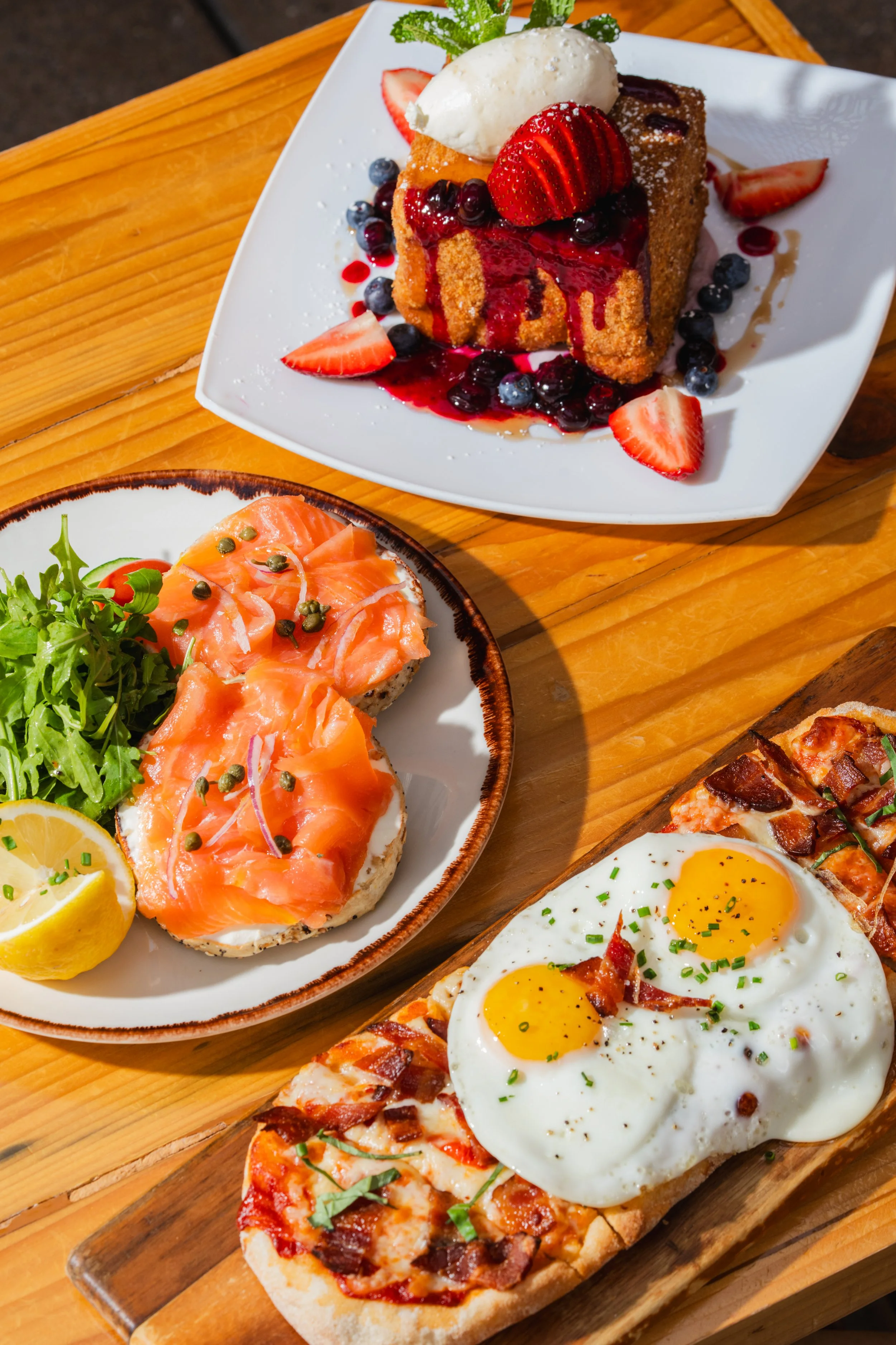 A dessert plate with a slice of cake topped with strawberries, blueberries, and ice cream, drizzled with berry sauce. Beside it, a plate with smoked salmon on bread garnished with capers and lemon. In the foreground, a pizza with sunny-side-up eggs, 