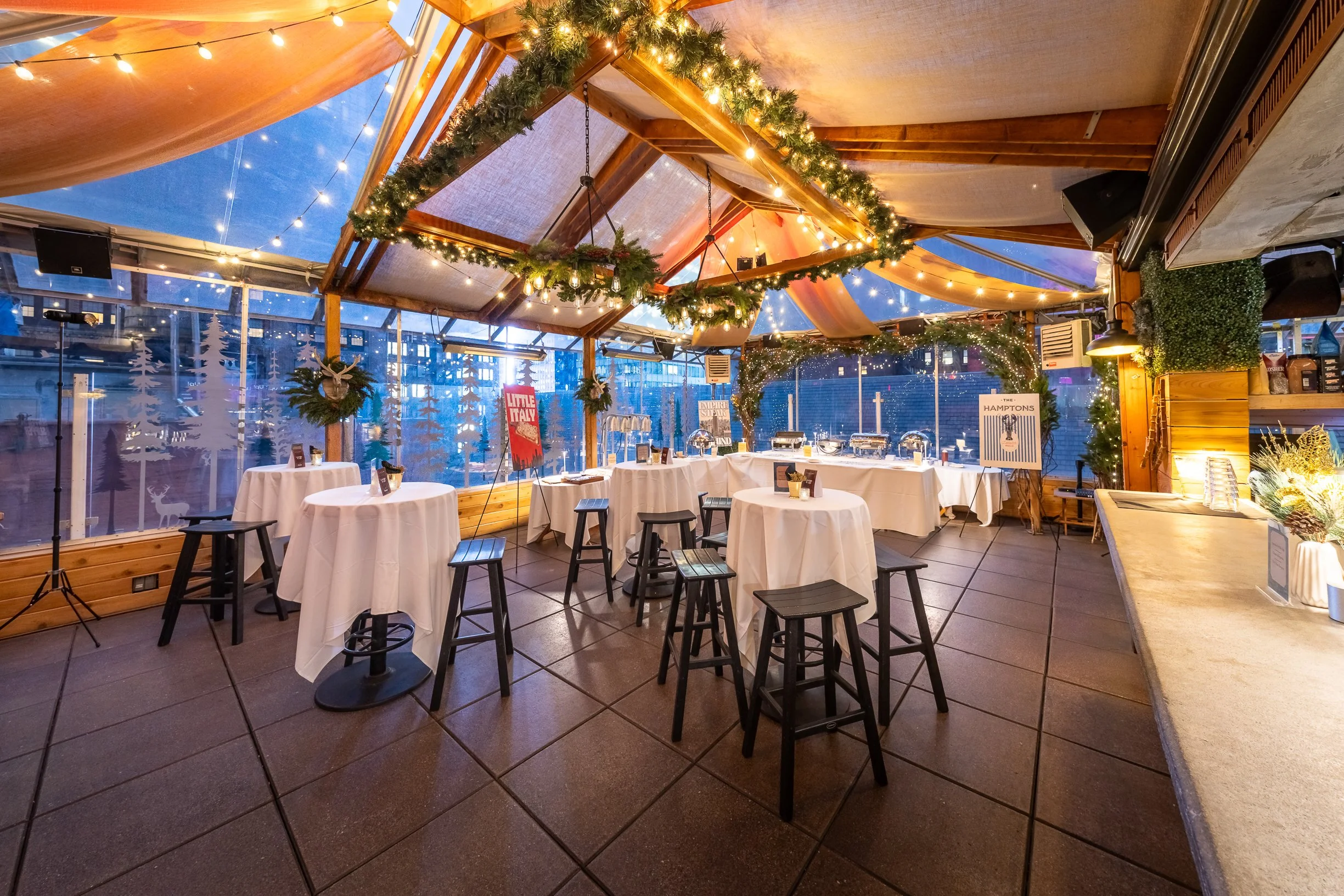 A decorated indoor party or event space with tables covered in white tablecloths, black stools, and string lights hanging from the ceiling. The space has wooden beams, garland with lights and ornaments, and large windows with a view of buildings outs
