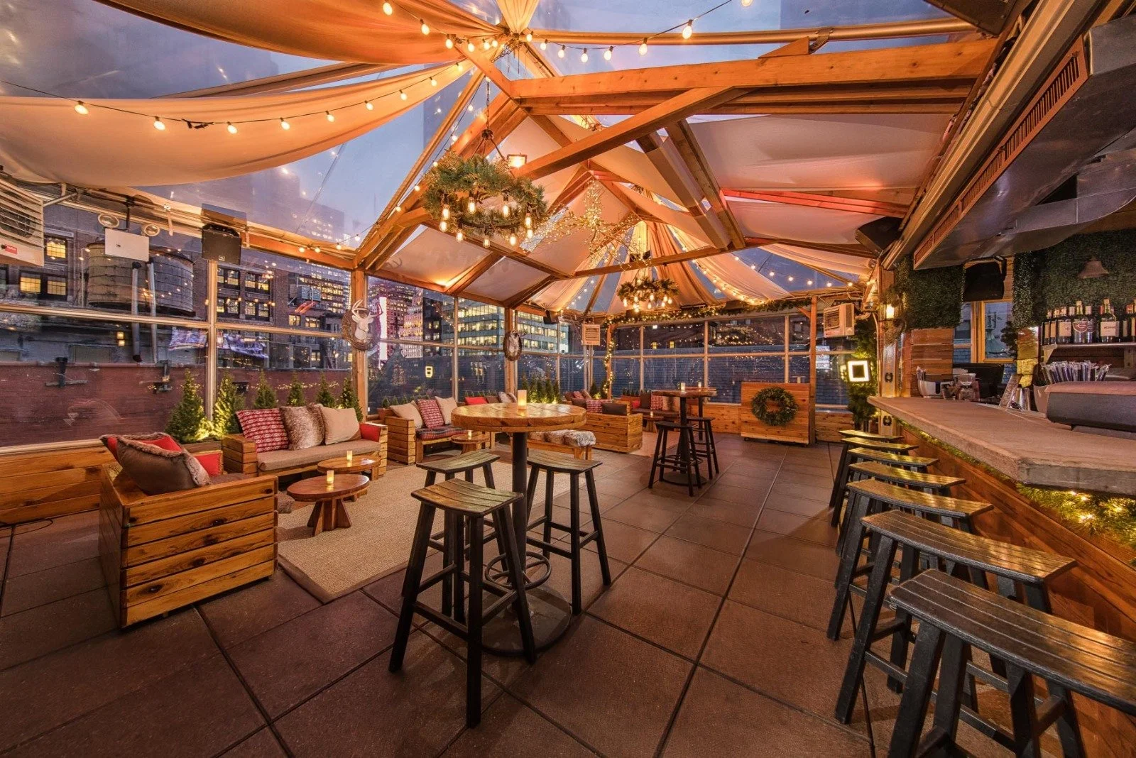 Cozy rooftop bar decorated with string lights and Christmas wreaths, featuring wooden furniture, cushioned seating, and a bar counter in the background, overlooking a city skyline at dusk.