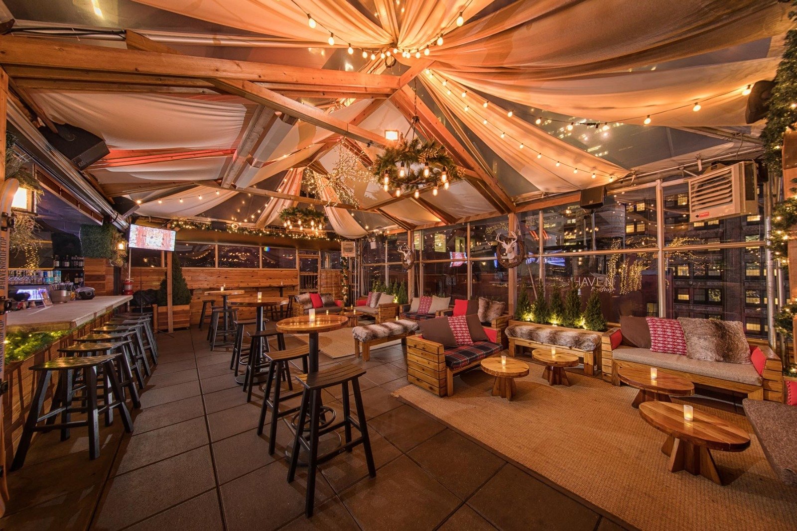 Interior of a cozy, rustic rooftop lounge decorated with string lights, wooden furniture, and holiday wreaths, with city buildings visible through large windows.