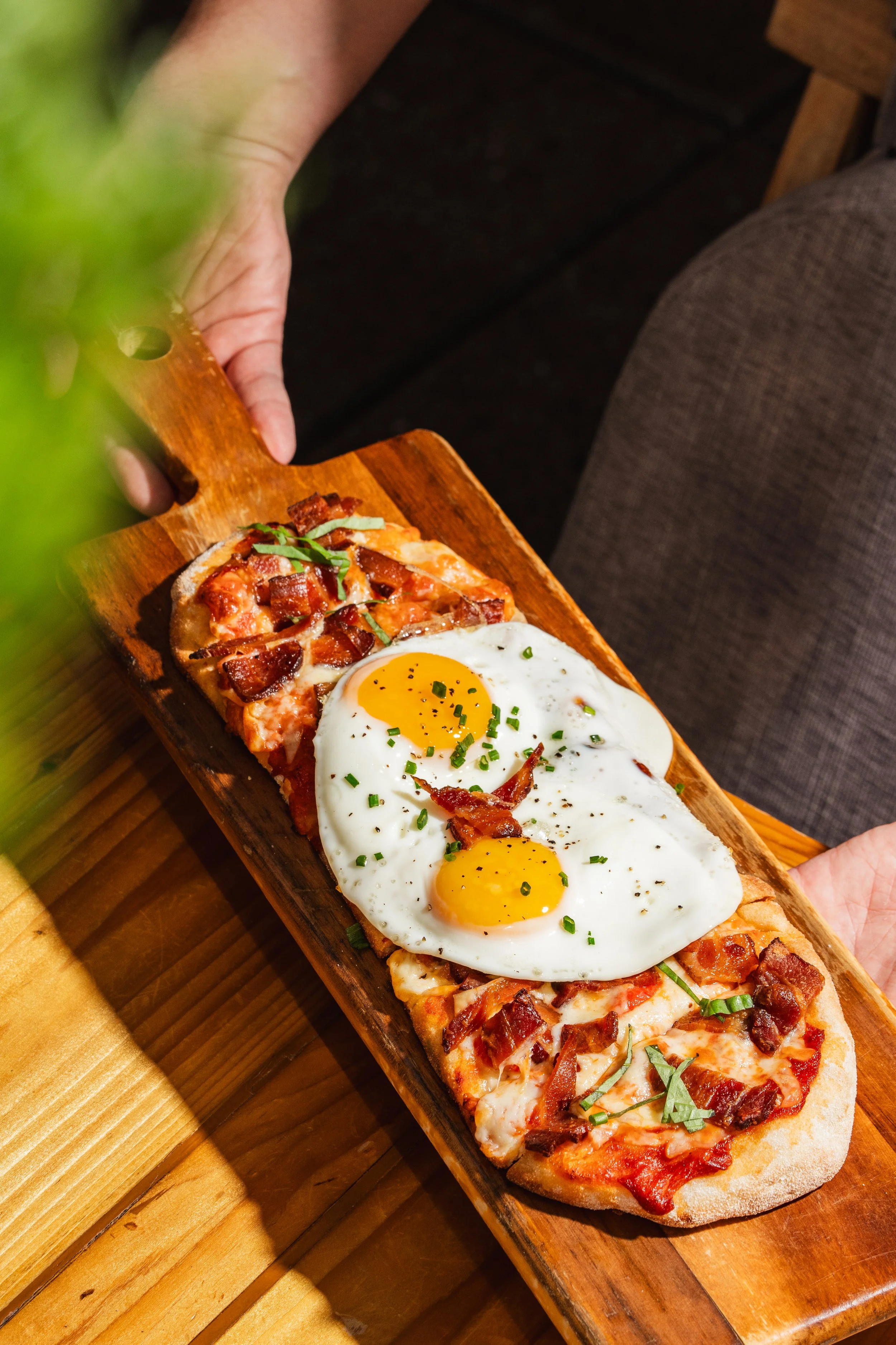 A wooden serving board with three slices of pizza topped with cheese, bacon, and green herbs, with two sunny side up eggs on top, garnished with chopped chives.