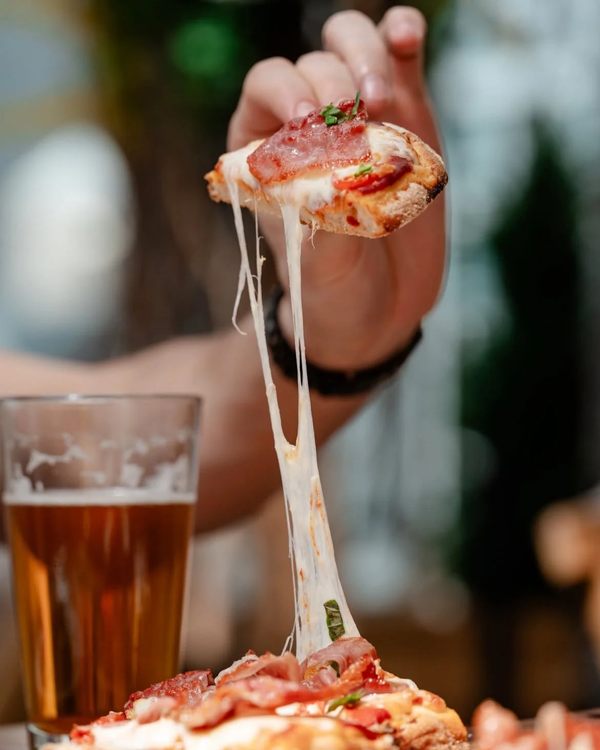 Person lifting a slice of pepperoni pizza with cheese stretching from the pizza to the slice, with a glass of beer in the background.