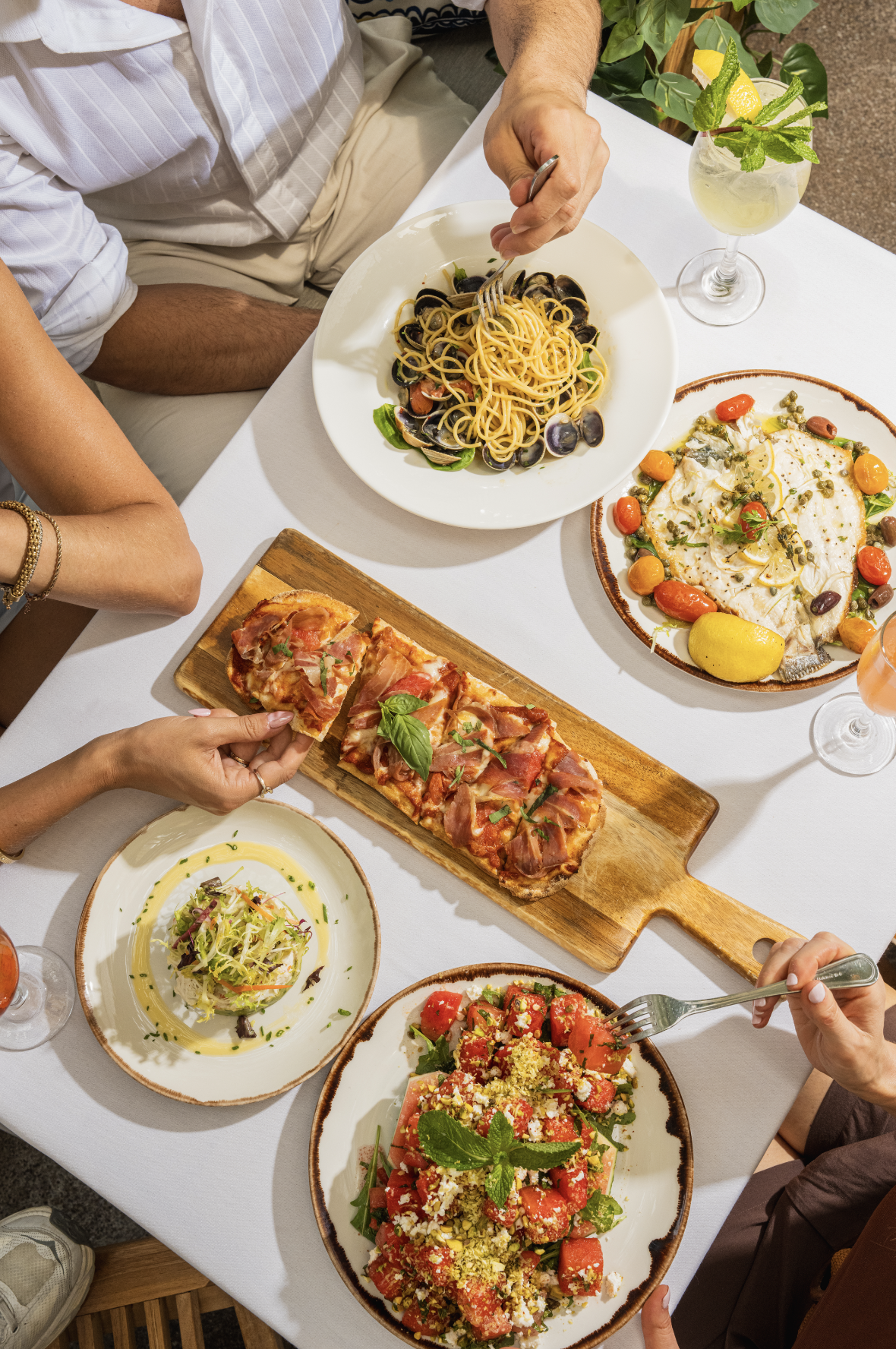 A table with various dishes including pasta with clams, pizza with prosciutto, and salads, along with drinks, in a dining setting.