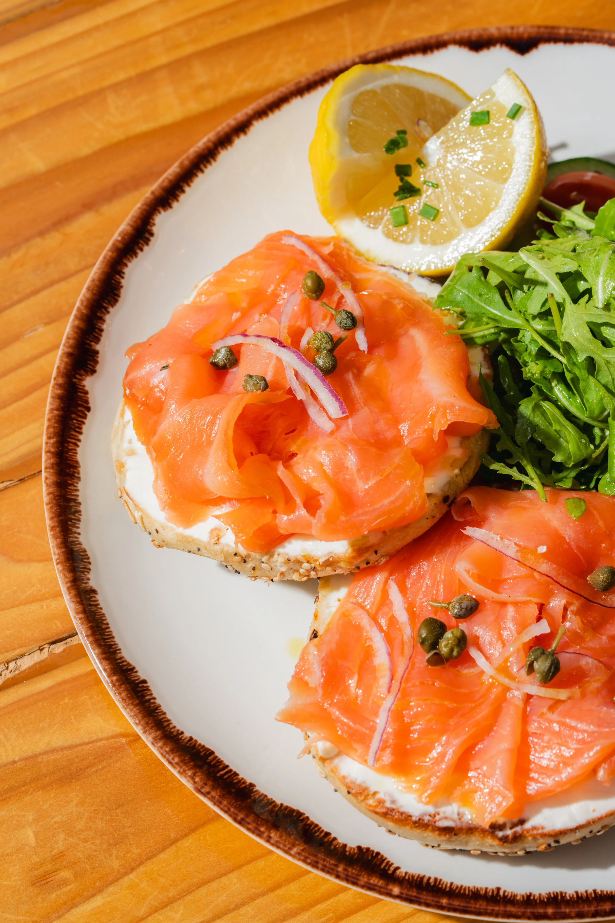 Close-up of smoked salmon on toasted bagel with cream cheese, garnished with capers, red onion, and parsley, with lemon slices and greens on the side, served on a white plate with a brown rim.
