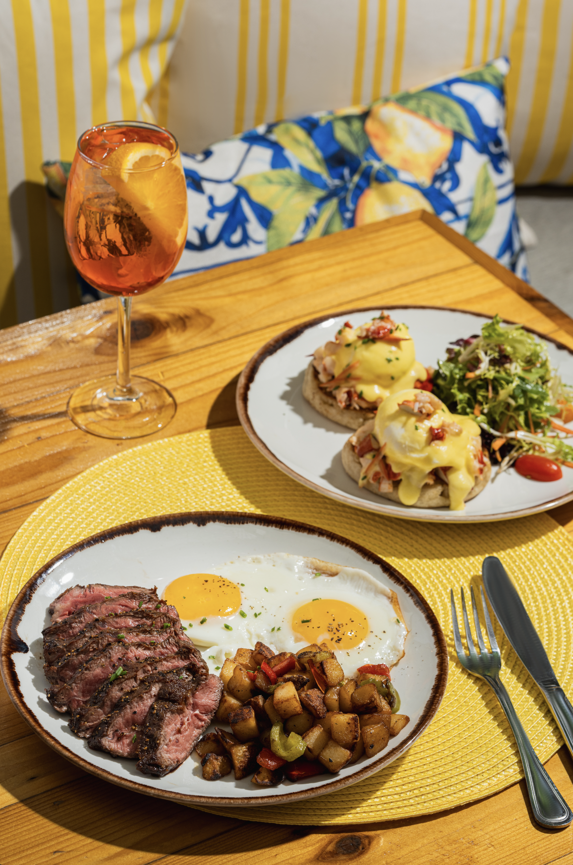 A breakfast plate with two sunny-side-up eggs, sliced steak, and diced potatoes, served on a ceramic plate. In the background, a plate with eggs Benedict and a side salad, along with a glass of orange-colored beverage with lemon slices, on a wooden t