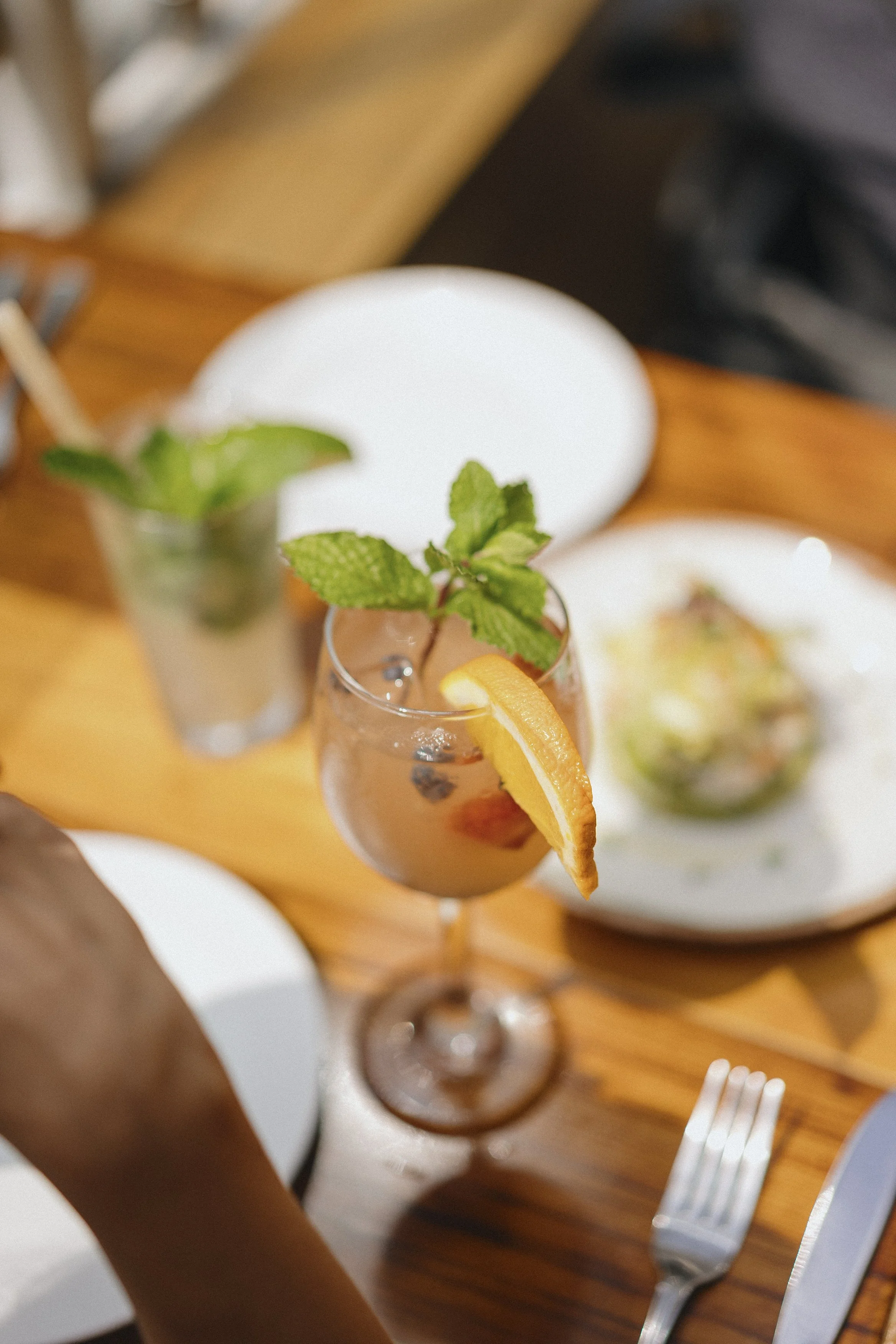 Close-up of a table with cocktails decorated with mint leaves and orange slices, with plates of food and utensils in the background.