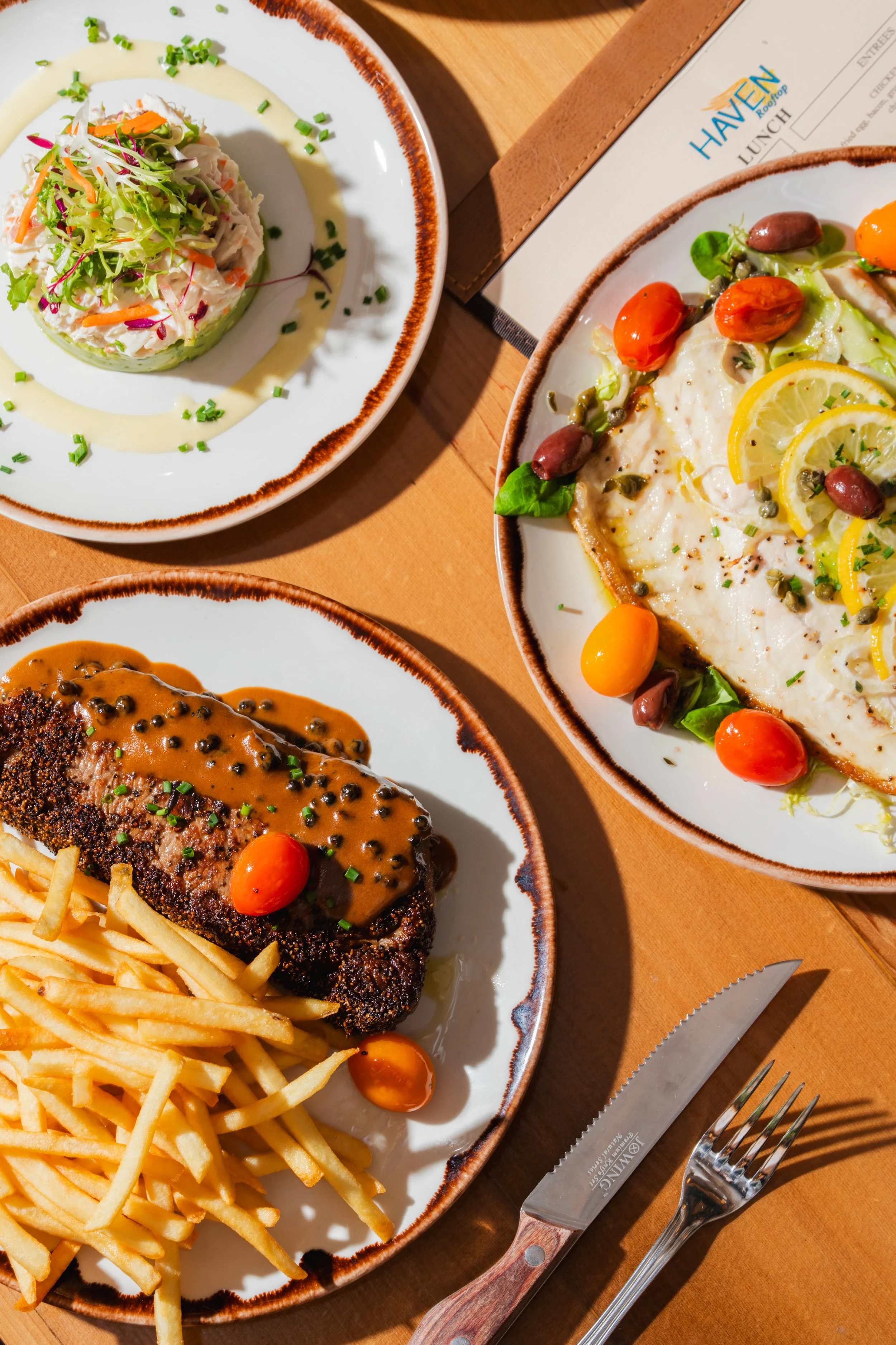 Close-up of a meal at a restaurant featuring a baked fish fillet with sauce, French fries, and cherry tomatoes, along with a salad with shredded vegetables, lemon slices, olives, and greens on ceramic plates on a wooden table.