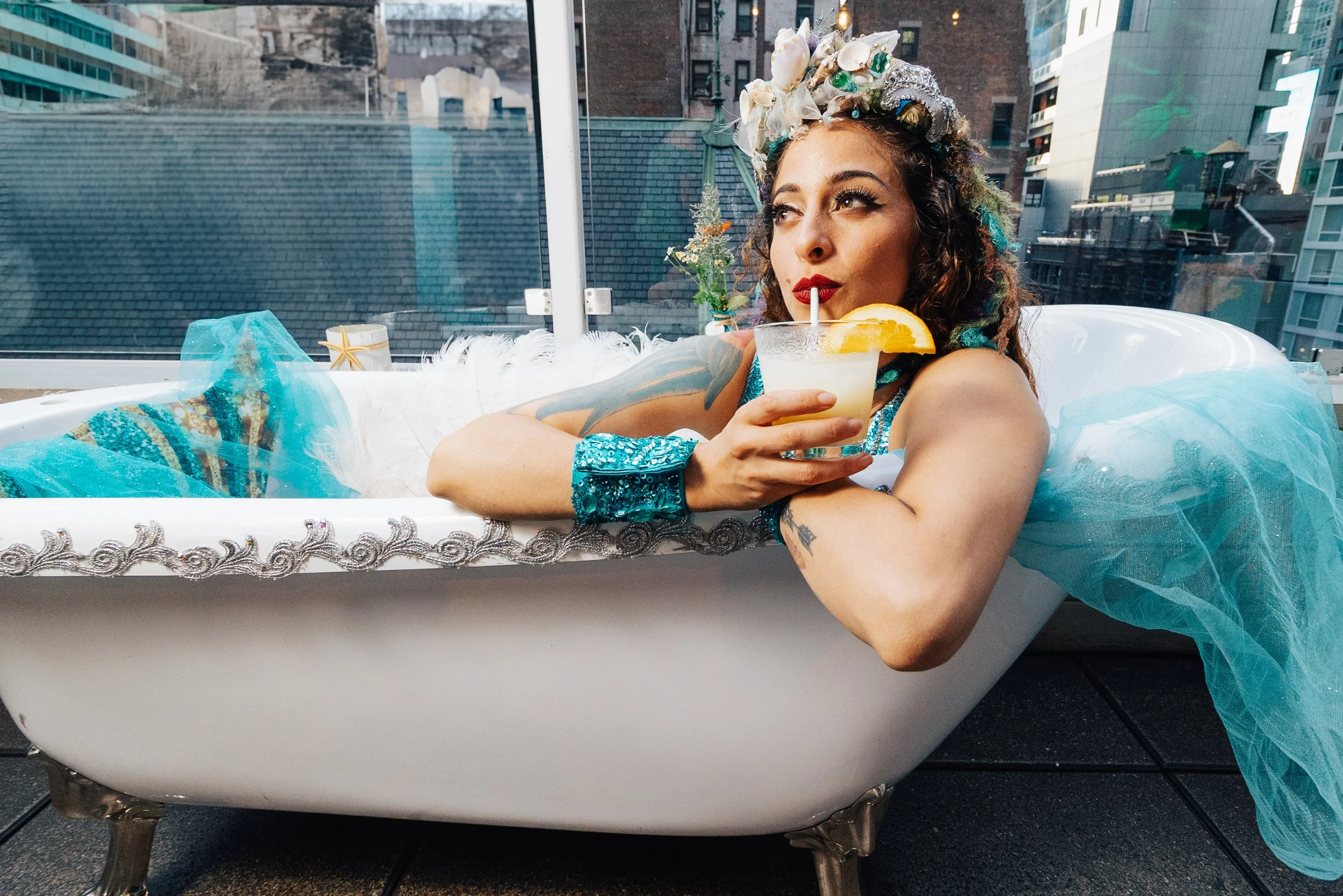 A woman with curly hair, wearing a floral headpiece, sitting in a vintage bathtub outdoors in an urban setting, enjoying a drink garnished with an orange slice.