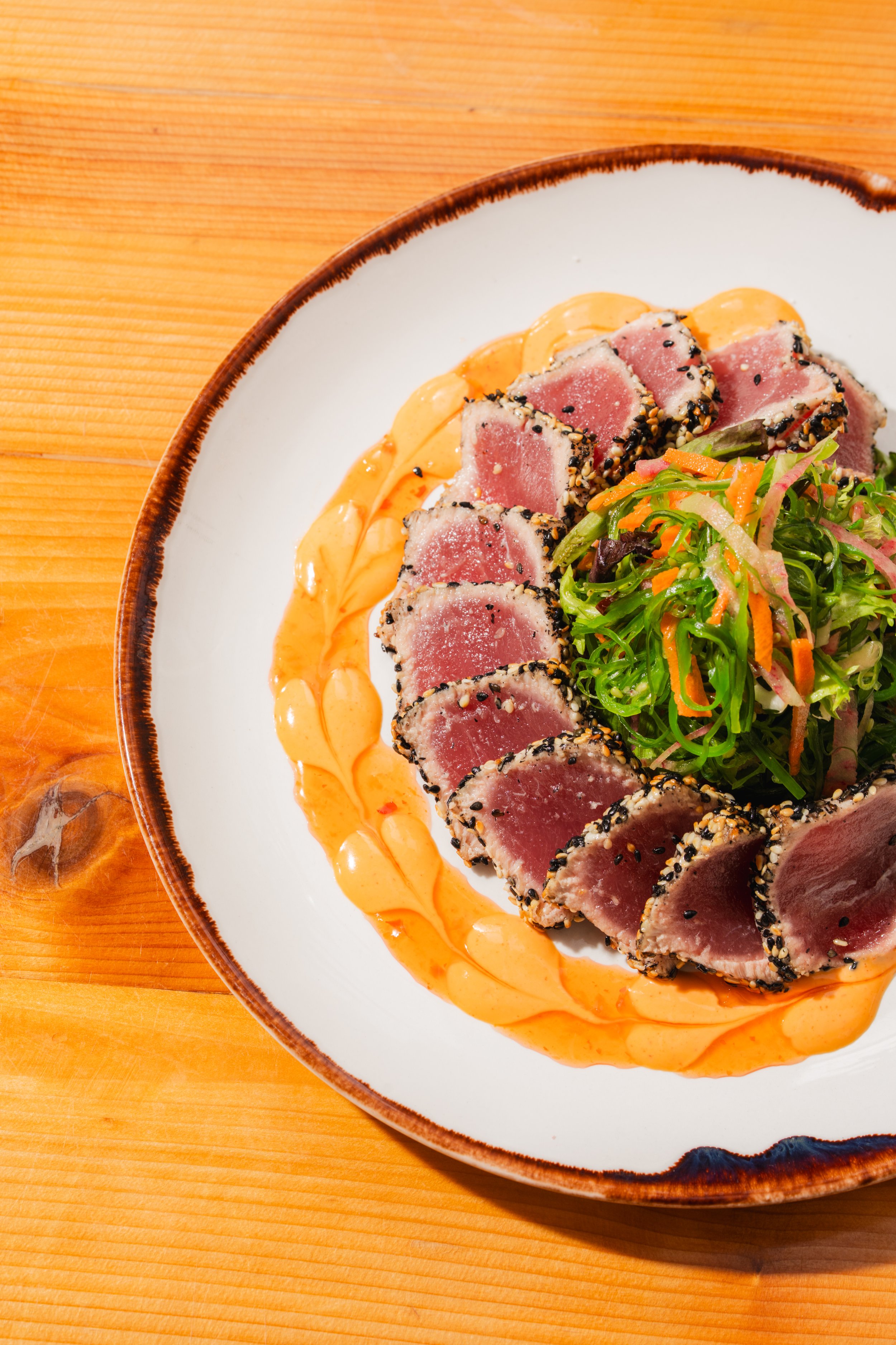 Slices of seared tuna with black sesame seeds served with a small salad of mixed greens, shredded carrots, and radish, on a white plate with a drizzle of orange sauce, on a wooden table.