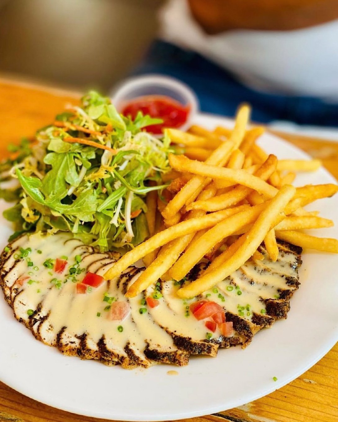 A plate with sliced grilled chicken topped with creamy sauce and chopped tomatoes, a side of French fries, a small salad with mixed greens, and a cup of ketchup.