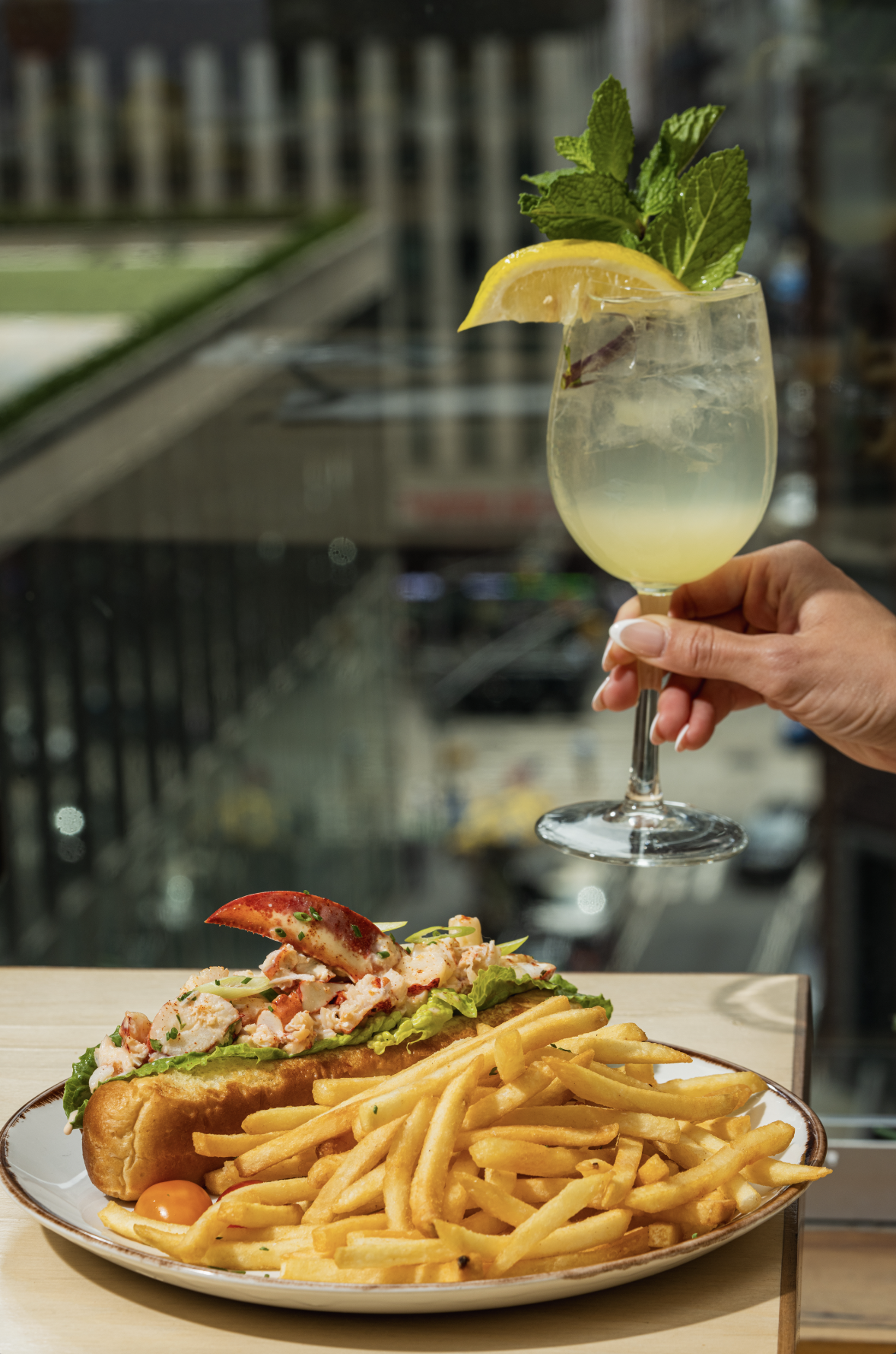 A hand holding a glass of lemonade with lemon wedge and mint, above a plate with lobster roll, French fries, and cherry tomatoes.