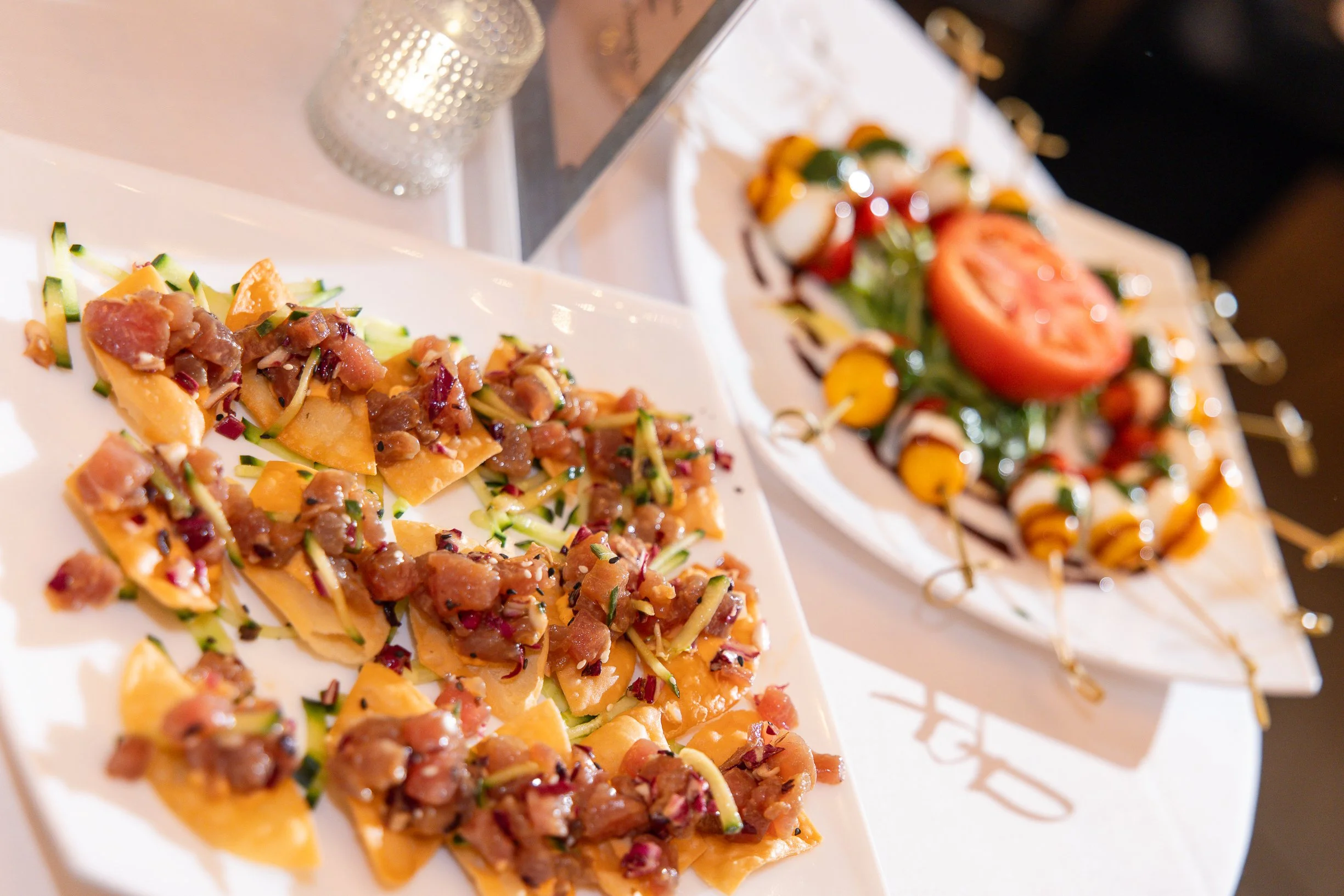 Close-up of two plates with appetizer items, including chopped meat and vegetables on crispy taco shells, and a skewer with tomato, cheese, and herbs, set on a white table with a glass of water in the background.