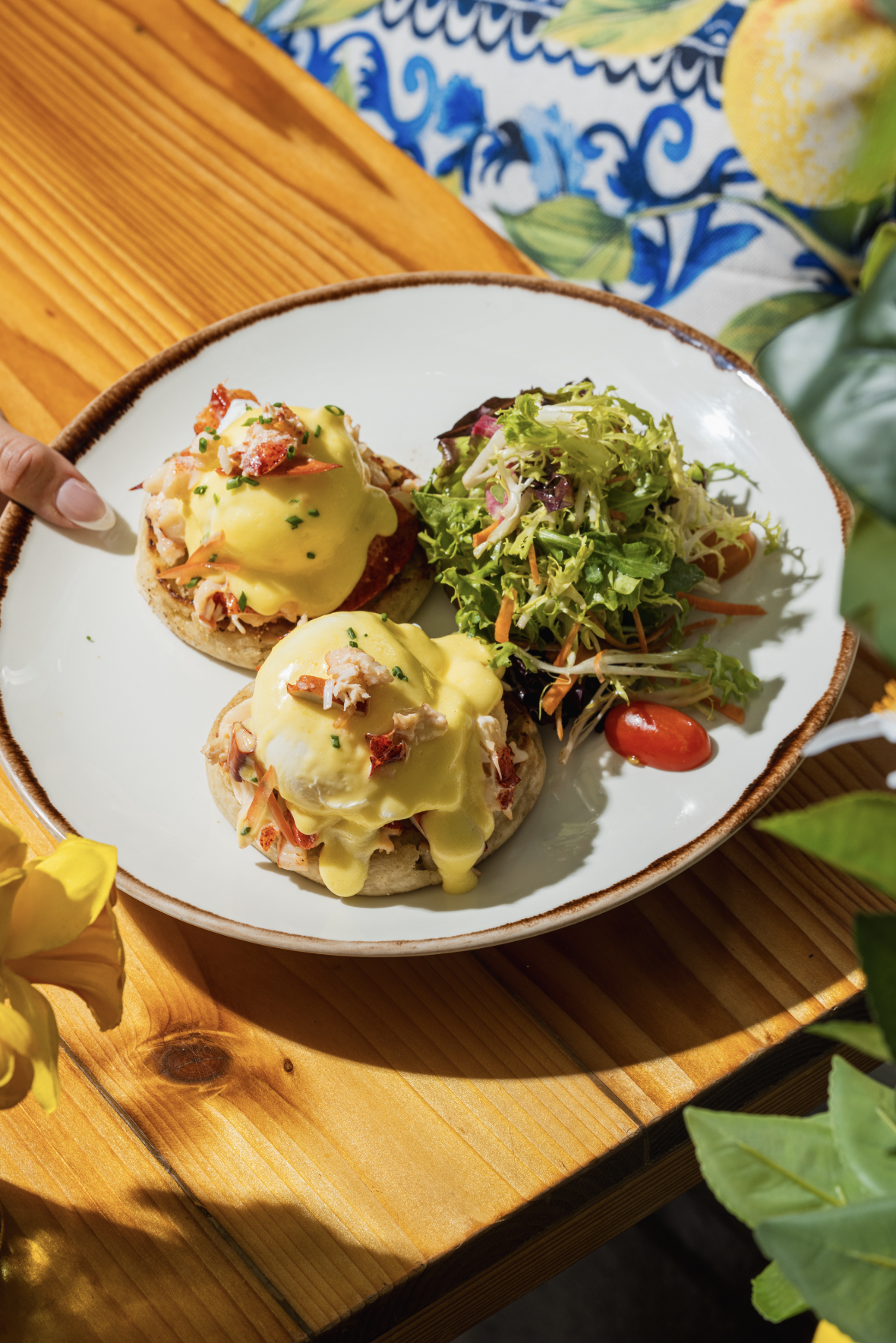 A plate with two eggs Benedict with hollandaise sauce and chopped chives, accompanied by a side salad with mixed greens, cherry tomatoes, shredded carrots, and shredded radishes on a wooden table.