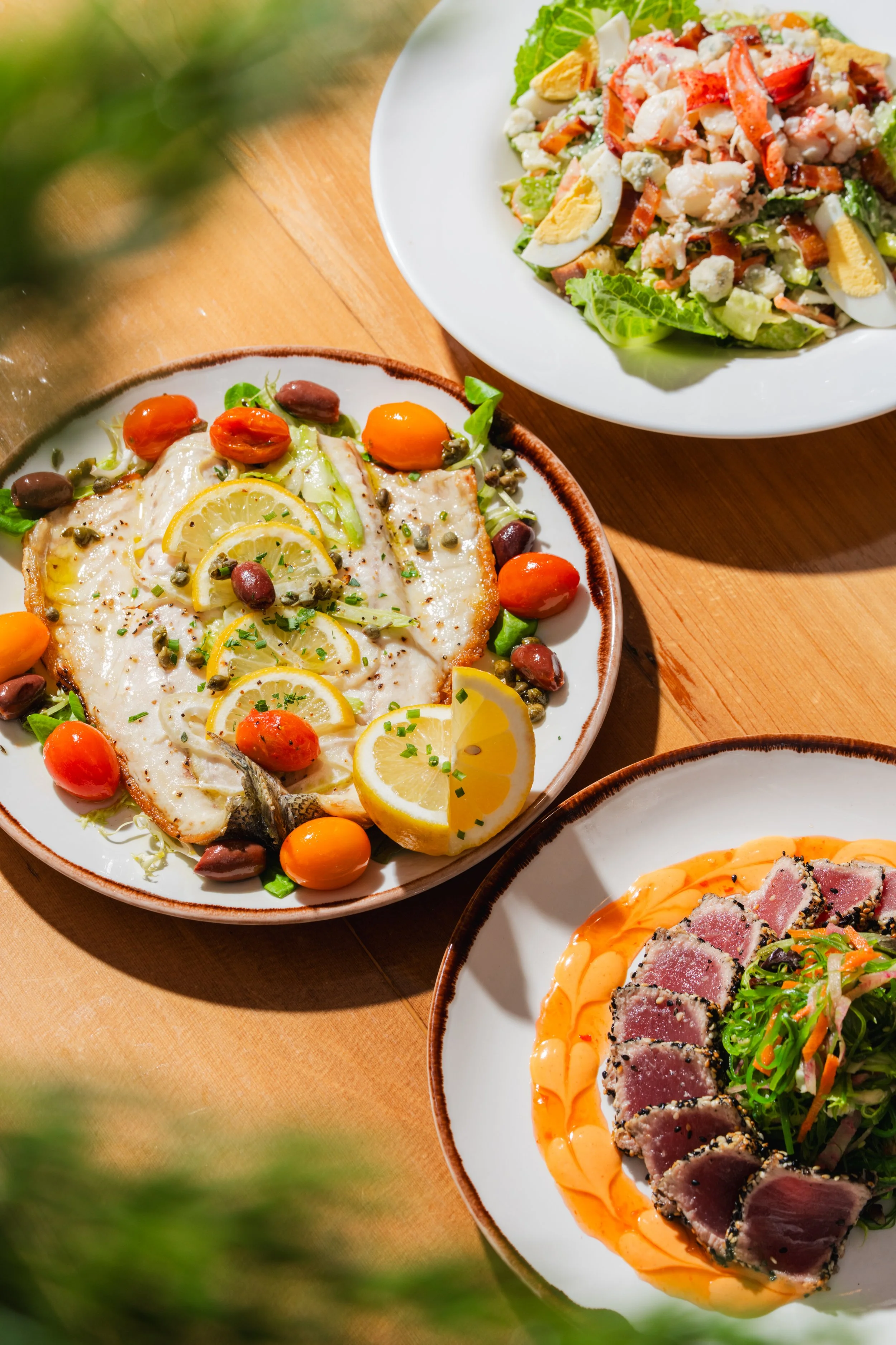 Three plates of food on a wooden table. The first plate has a fish dish garnished with lemon slices, capers, cherry tomatoes, and herbs. The second plate contains a salad with lettuce, hard-boiled eggs, crab meat, and bacon. The third plate, only par