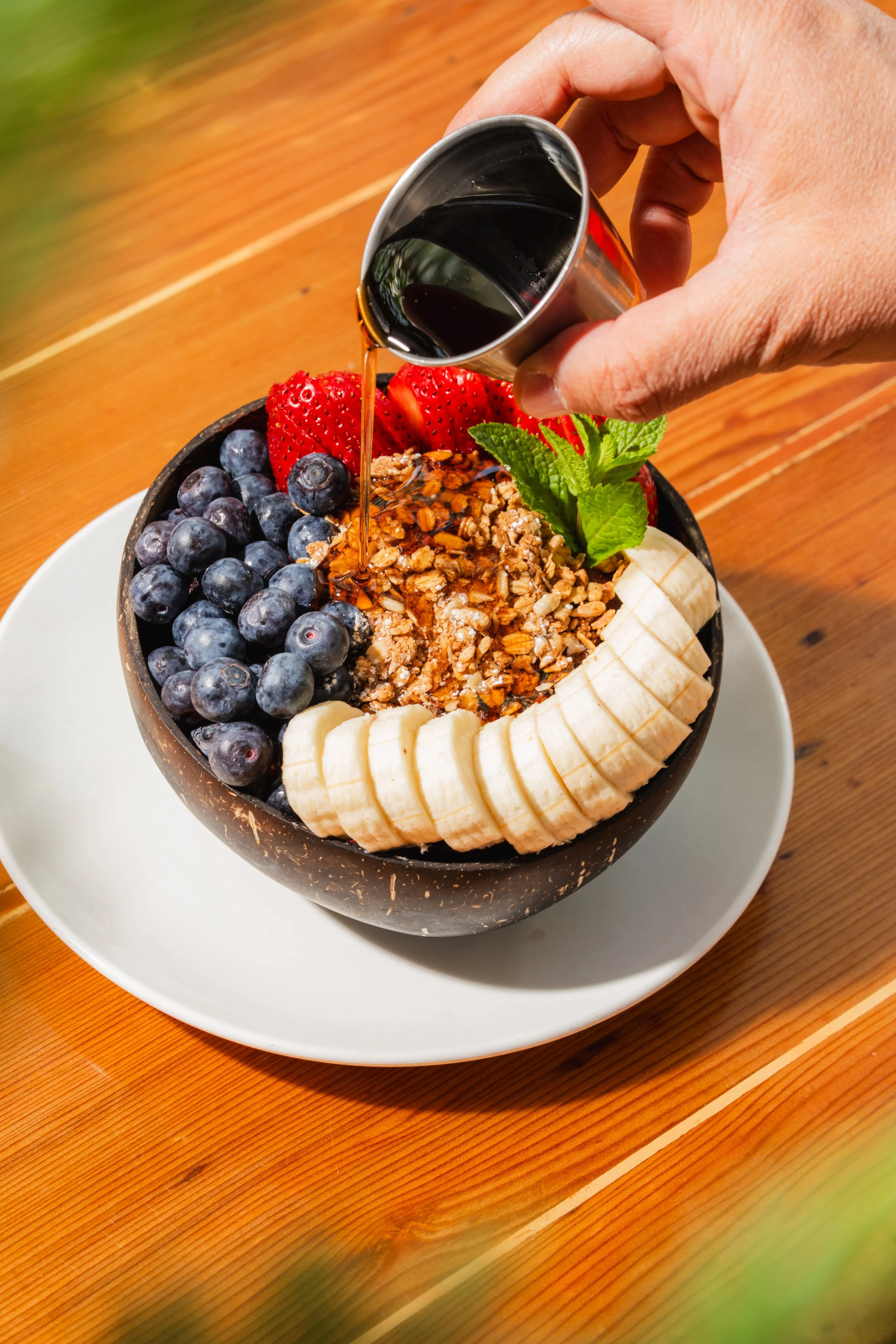 A bowl filled with blueberries, strawberries, sliced bananas, granola, and fresh mint leaves, with dark syrup being poured over the top.