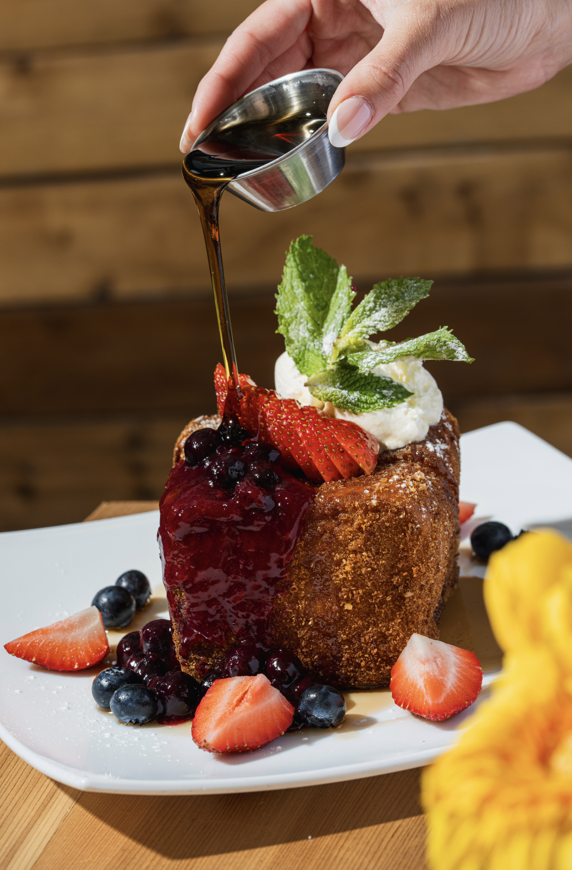 Deep-fried dessert topped with strawberries, blueberries, mint, whipped cream, and drizzled with syrup on a white plate.