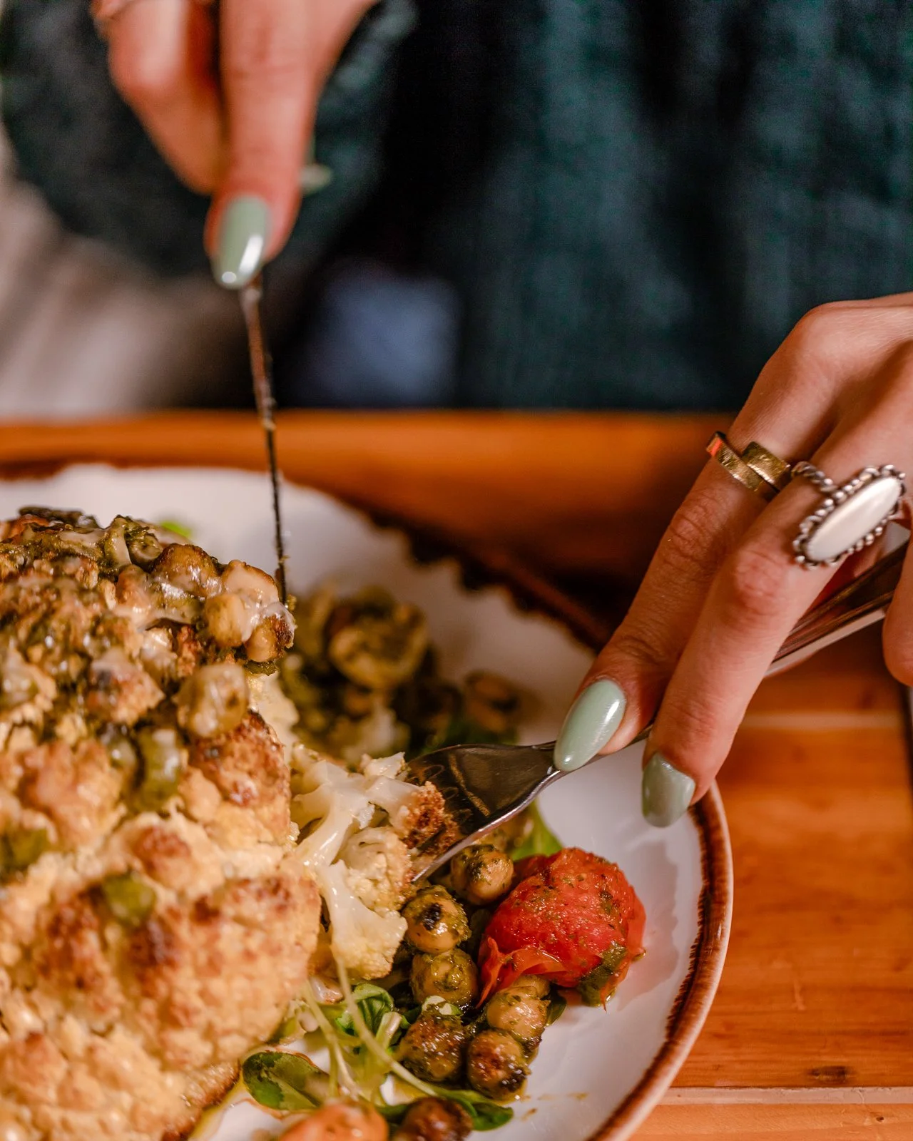 Person using a fork to serve a slice of baked casserole with cheese, peas, and possibly meat, on a decorative plate with roasted tomatoes and herbs.