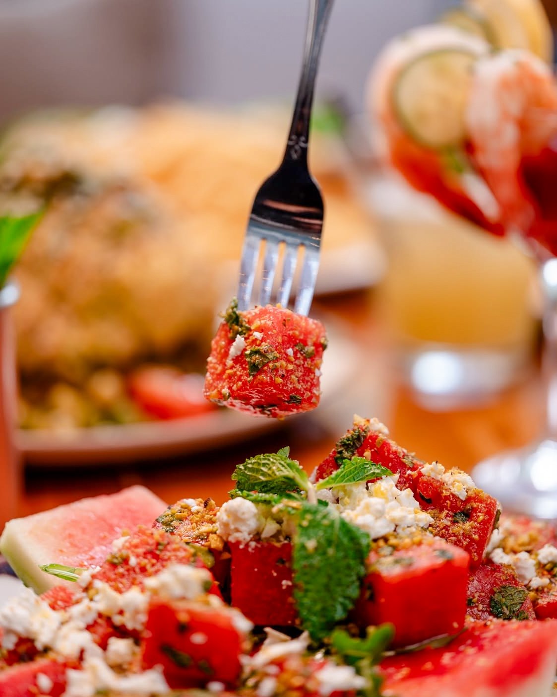 A fork holding a piece of watermelon salad topped with herbs and cheese, above a plate of watermelon cubes garnished with mint and cheese.