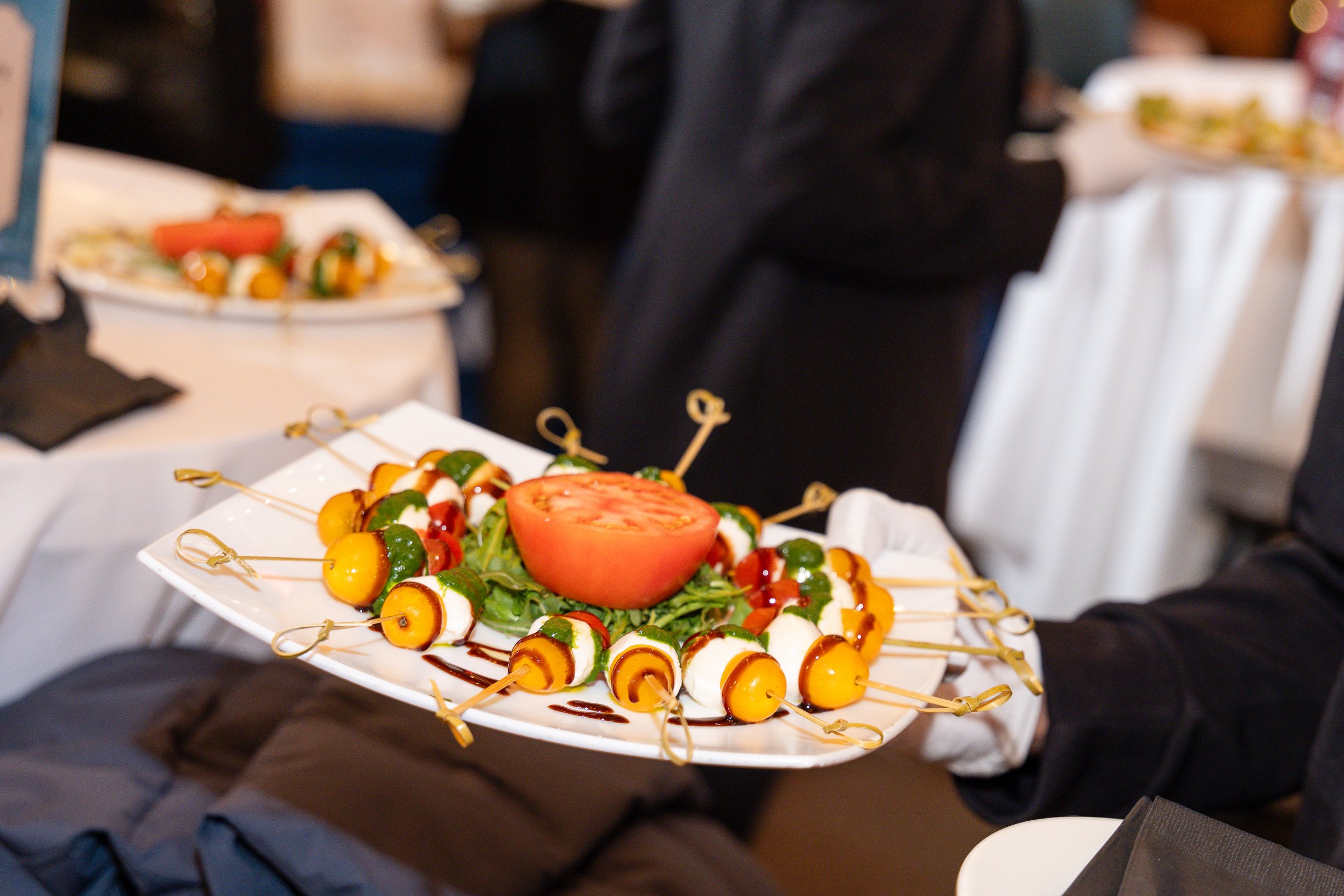 A white plate filled with colorful appetizer skewers, topped with a sliced tomato, and garnished with green herbs. The skewers include yellow, white, and red ingredients. The plate is held by a person dressed in black at a formal event or dinner.