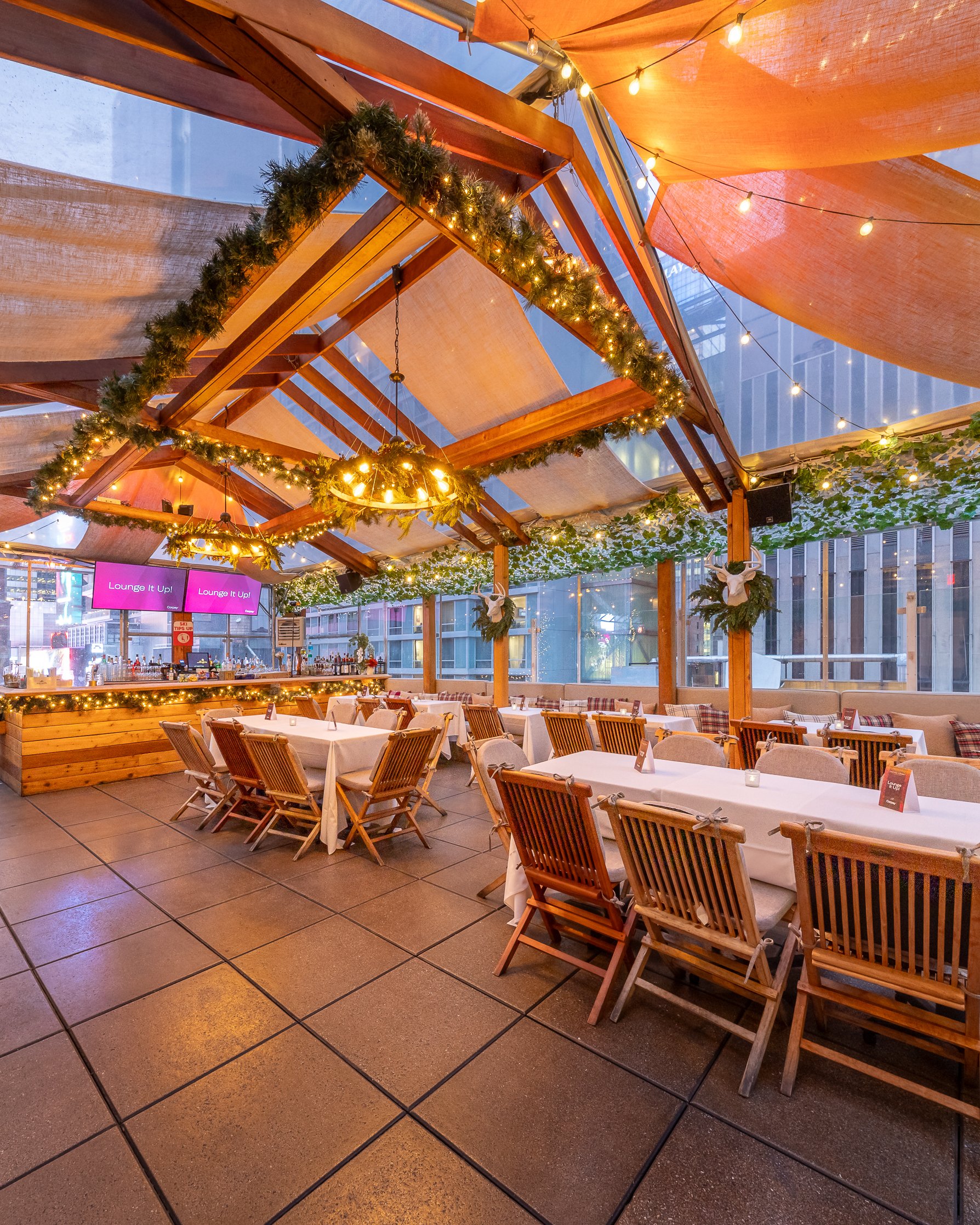 Empty outdoor restaurant patio with wooden chairs and tables covered in white tablecloths, decorated with string lights and garlands, with city buildings visible through glass windows.