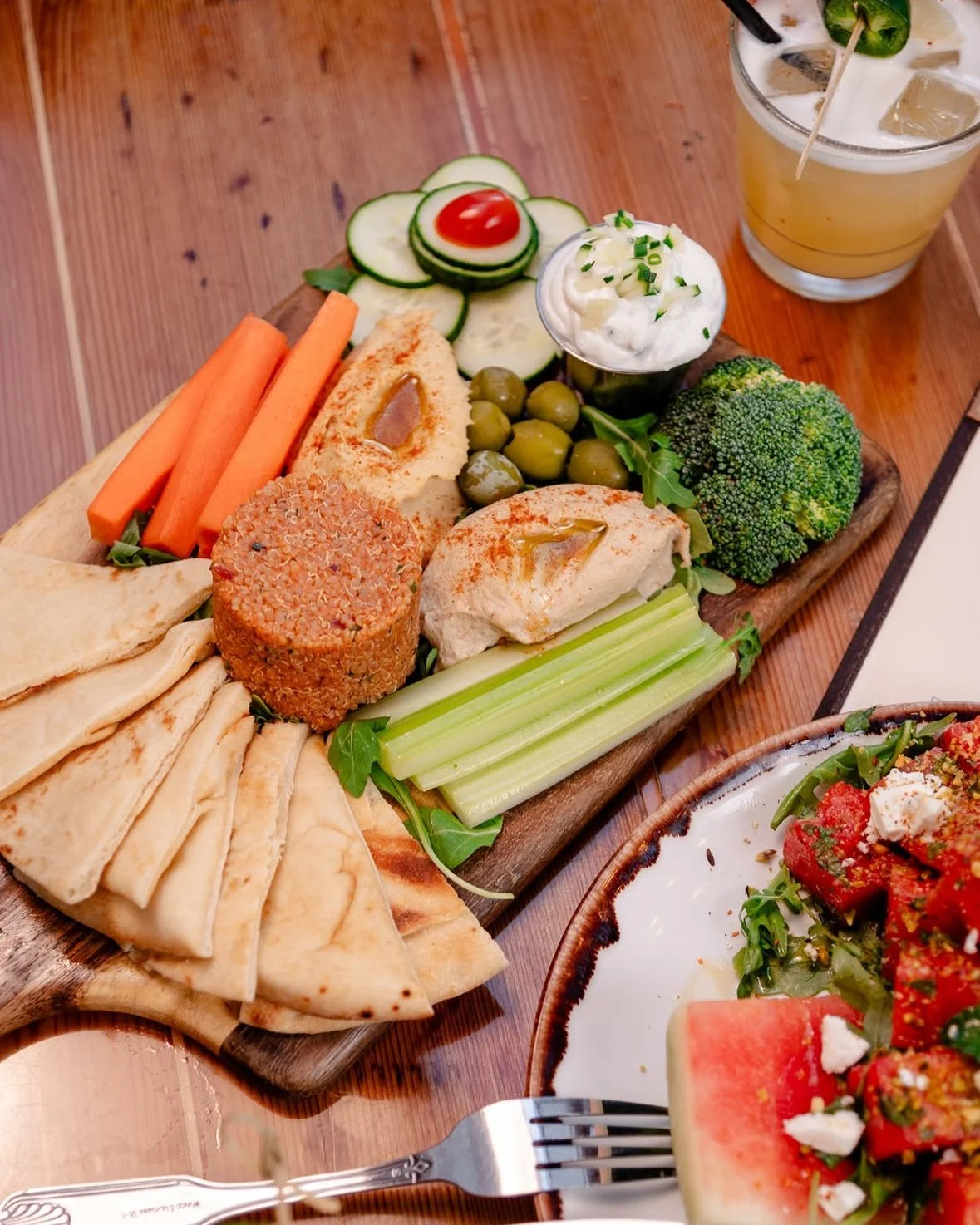 A wooden platter with assorted vegetables, hummus, and a small bowl of tzatziki sauce, accompanied by a glass of lemonade with lime, ice, and a straw, on a wooden table.